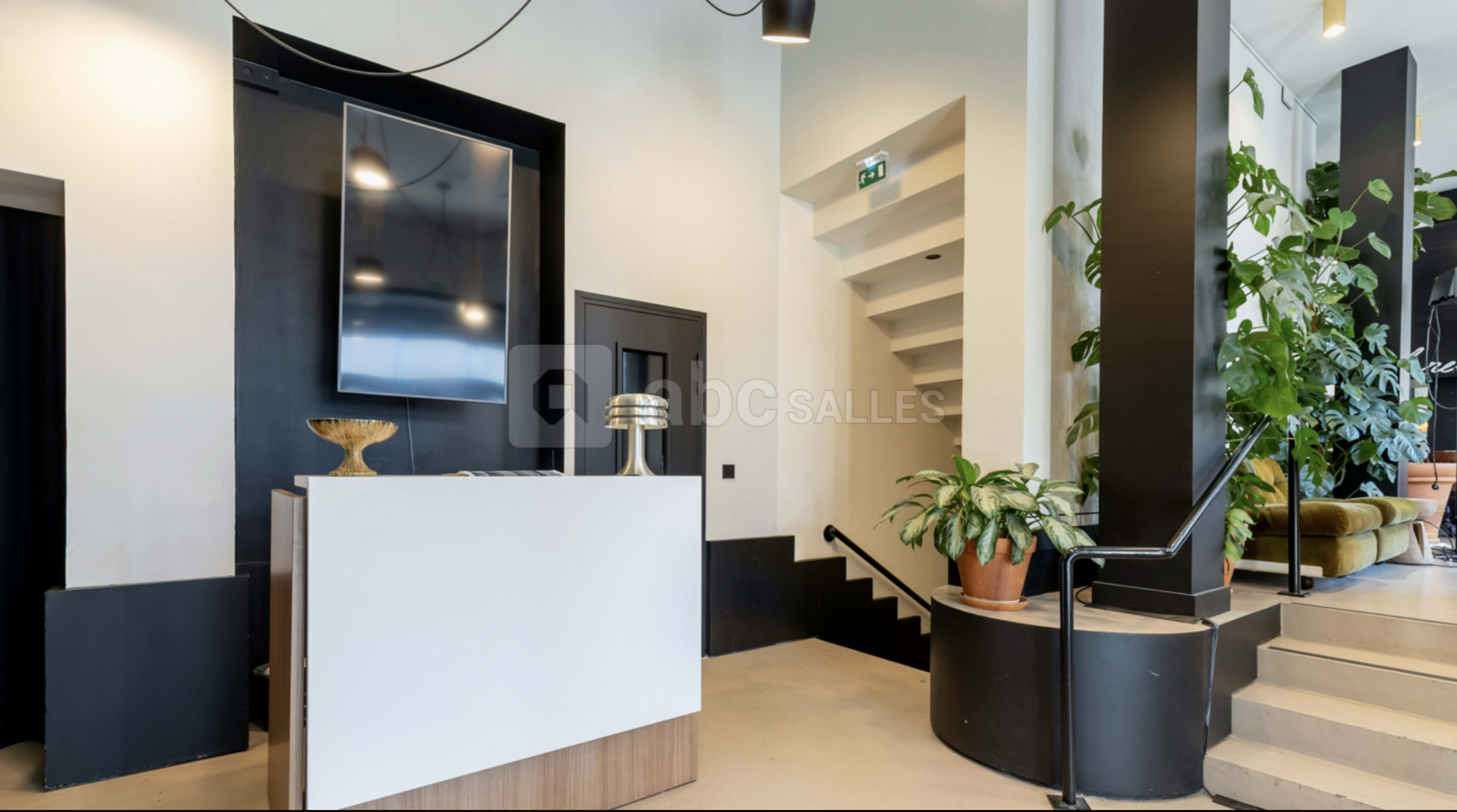 The image shows a modern reception area with a sleek front desk, a large television screen on the wall, and decorative plants.