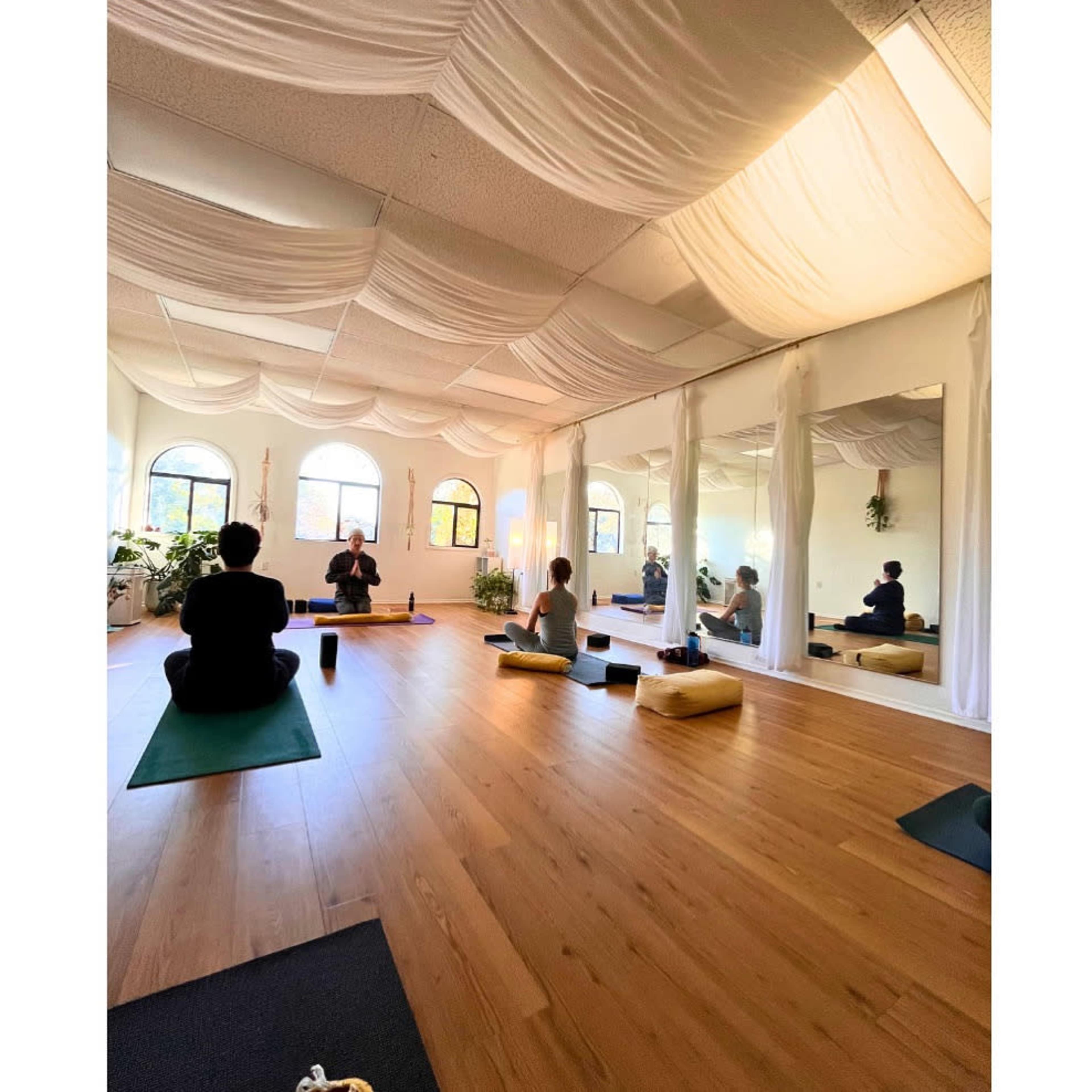 The image shows a serene yoga studio with wooden flooring, soft lighting, and mirrored walls, where individuals are engaged in a yoga practice.