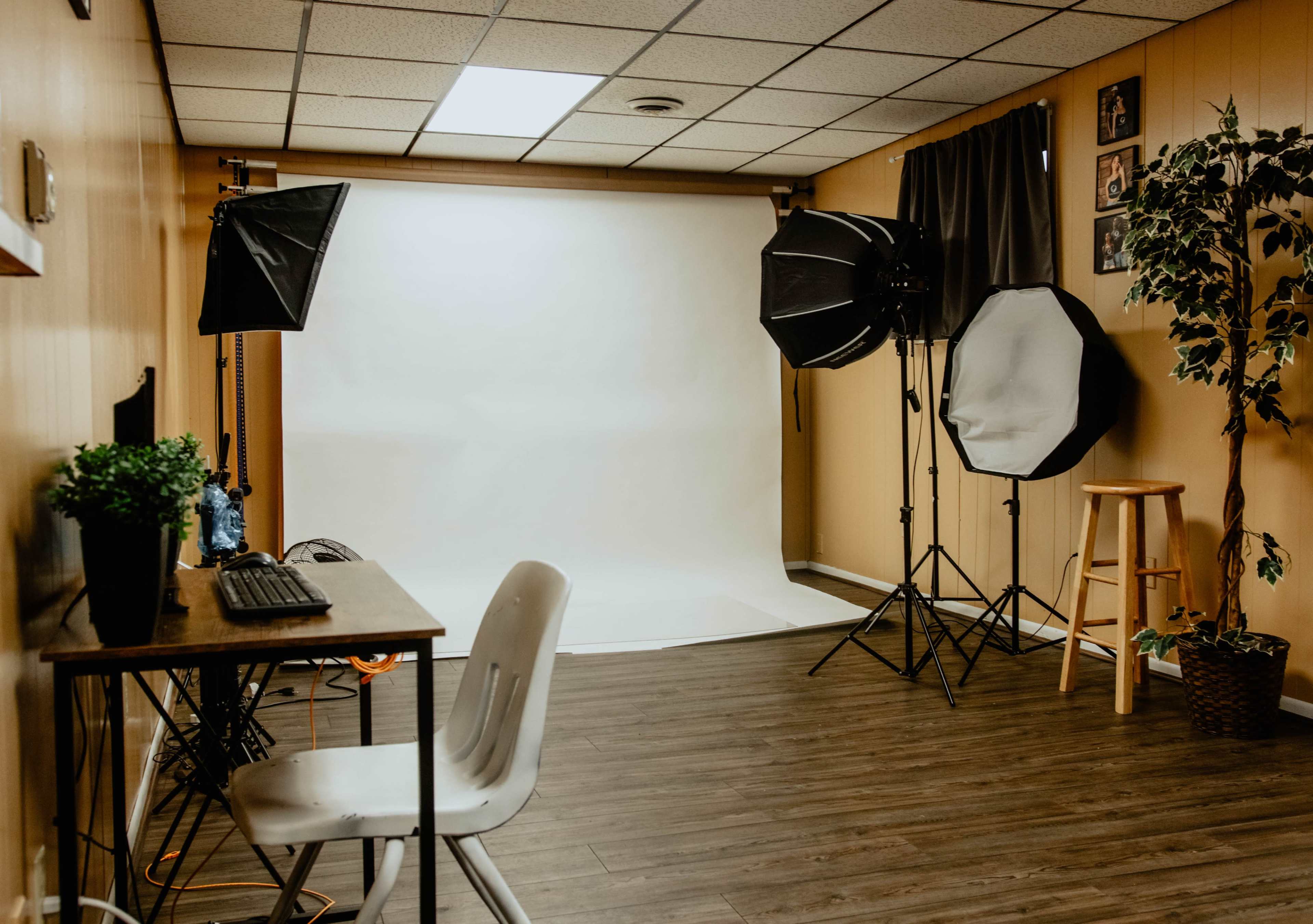 A small photography studio with a light backdrop, two softbox lights, a plant, a stool, and a desk with a computer chair.