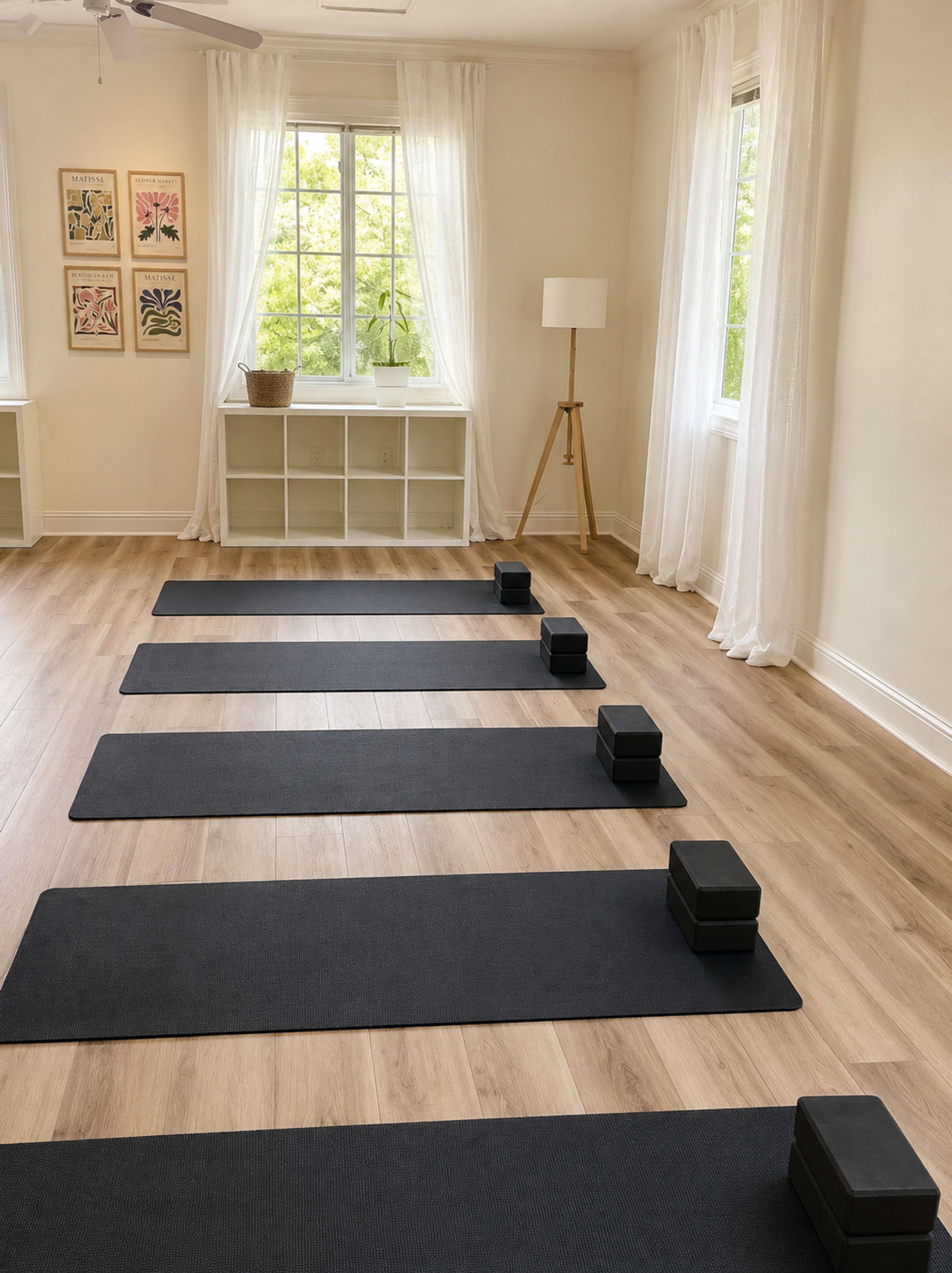 The image shows a bright and airy yoga studio featuring several black mats arranged in parallel lines, with foam blocks placed on top of each mat.