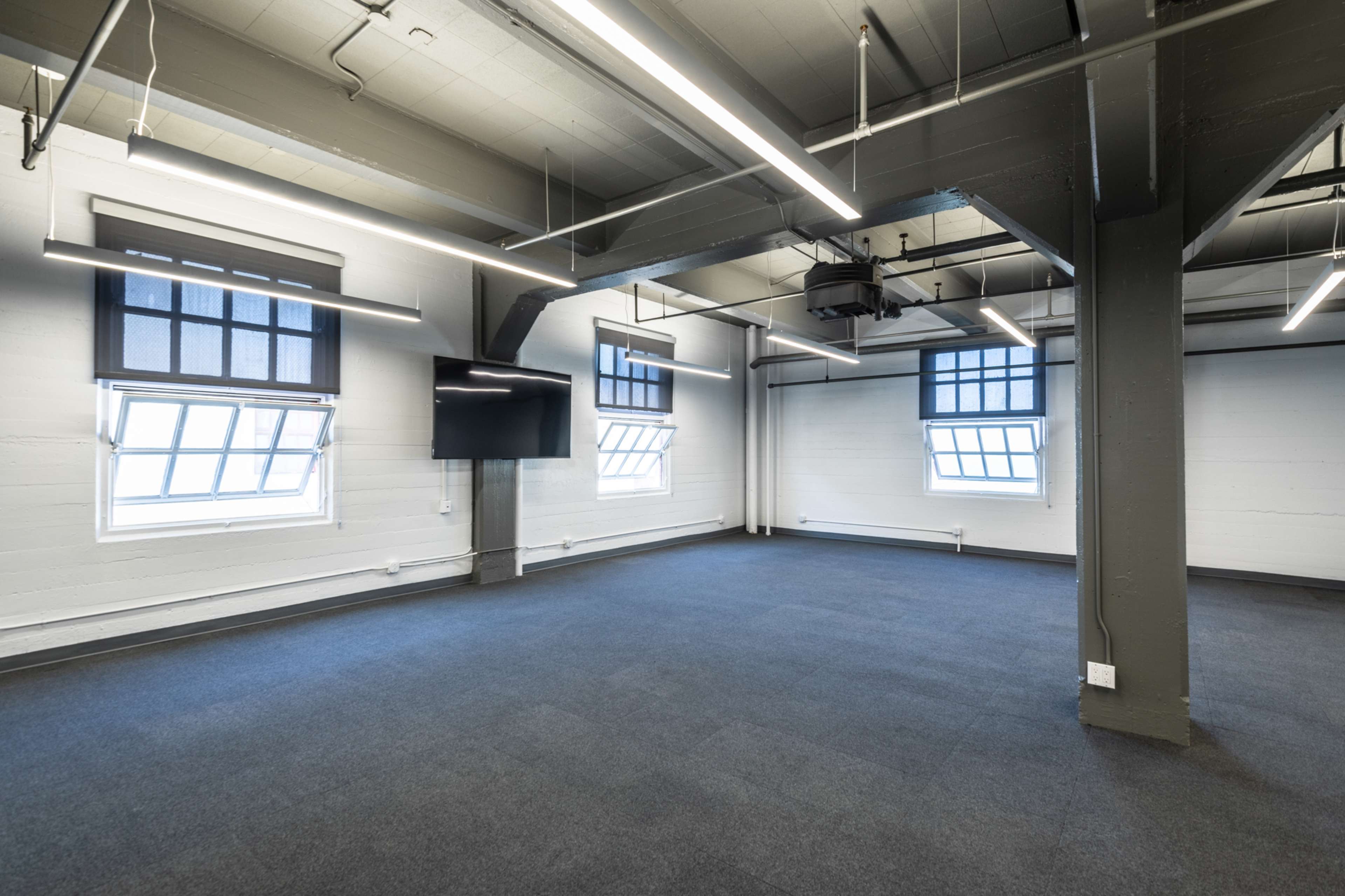 A spacious, empty room with gray walls, a dark carpeted floor, and large windows featuring blinds, illuminated by overhead lighting.