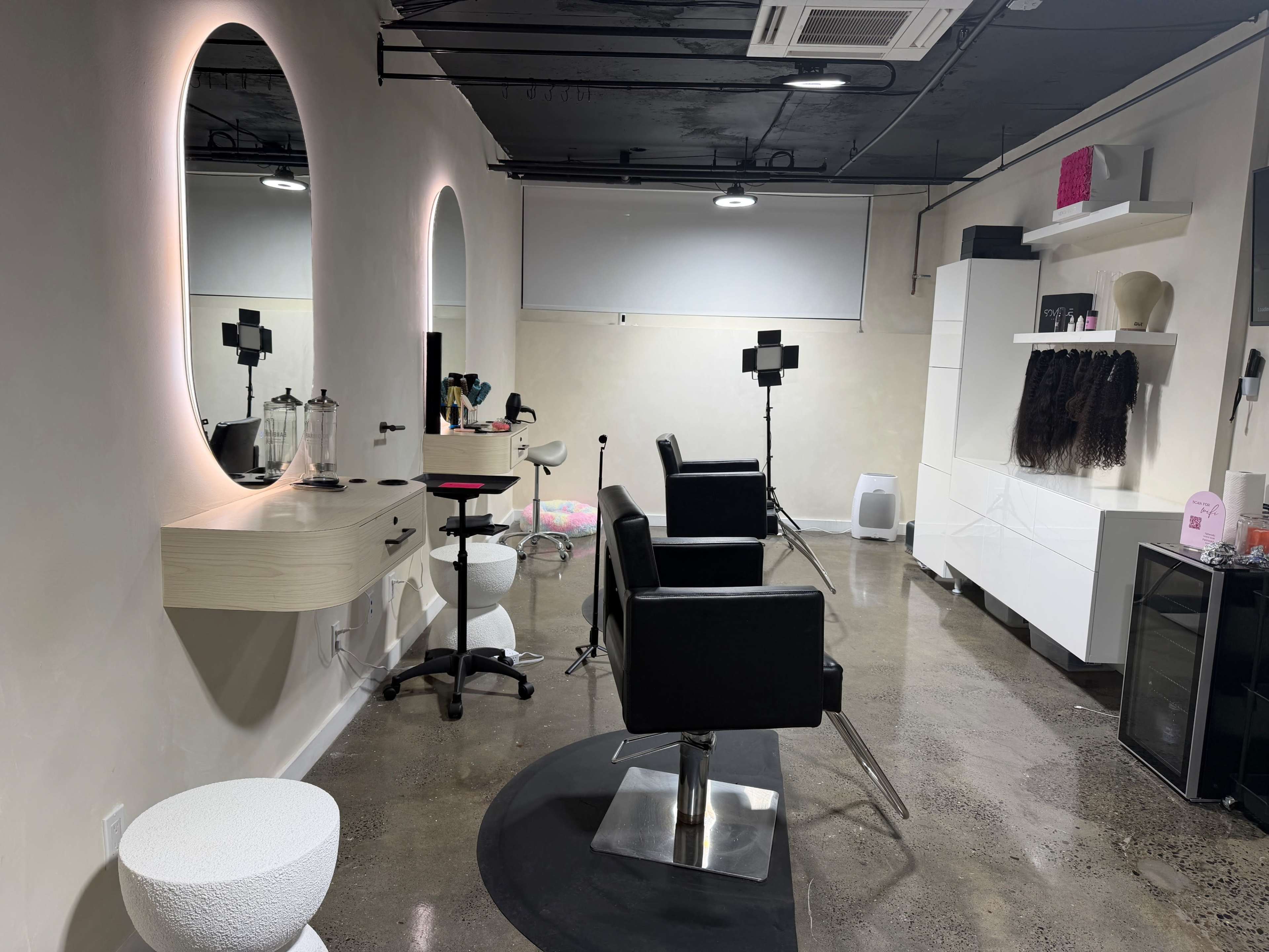 The image shows a modern hair salon interior featuring two black styling chairs, a mirror with ambient lighting, and a shelving unit displaying hair extensions and beauty products.