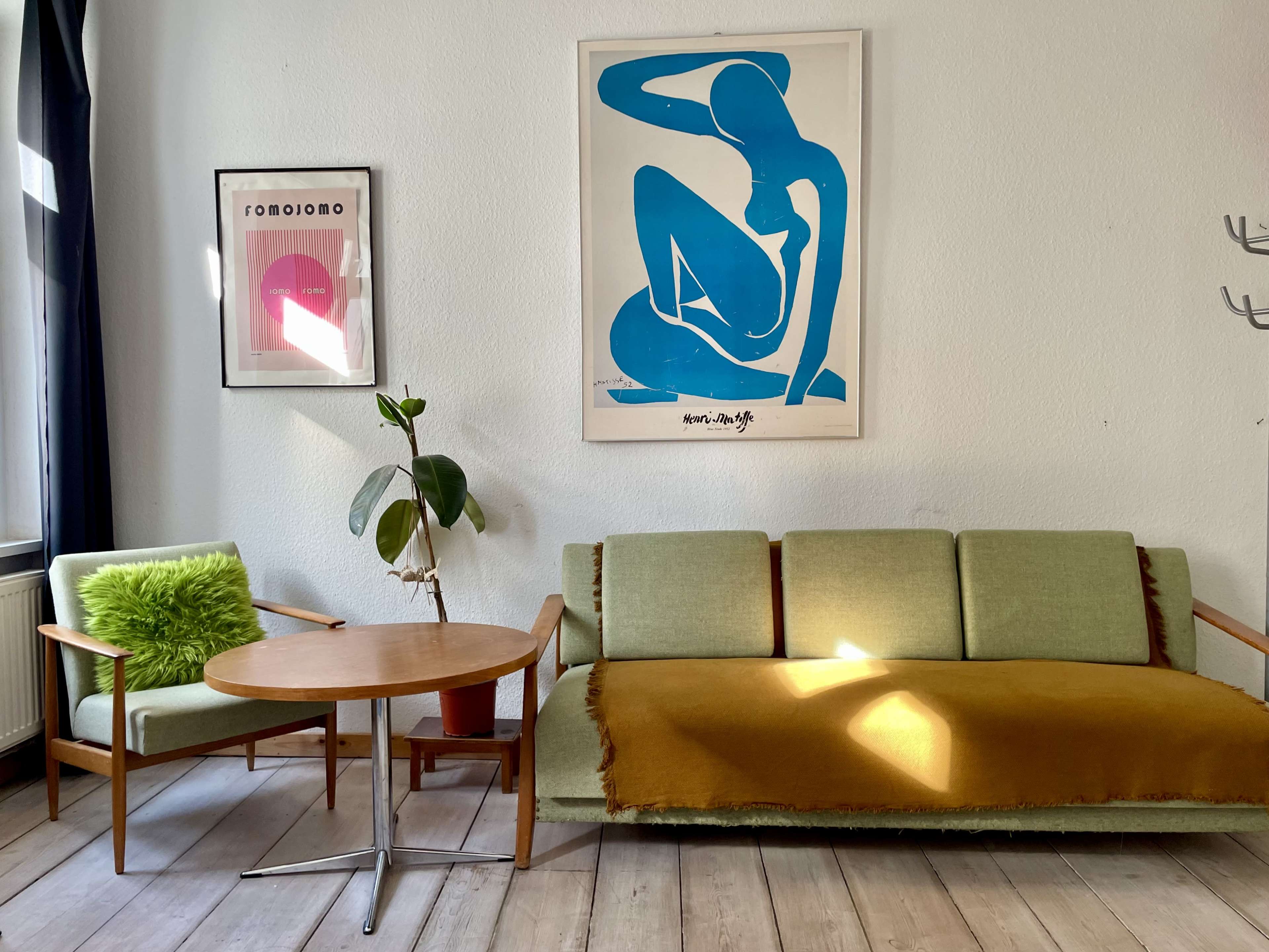 The image shows a living room with a green sofa, a round wooden table, and a green armchair adorned with a faux fur pillow, along with a framed poster on the wall.