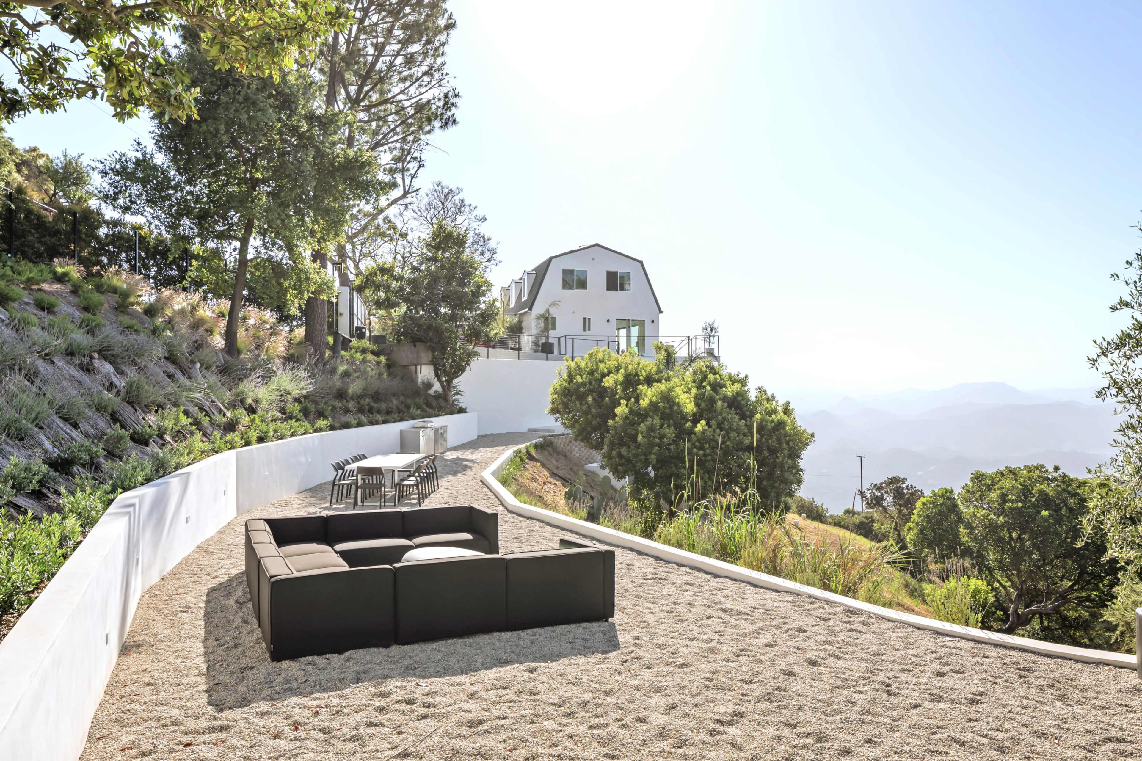 A modern house sits on a hillside surrounded by greenery, with a spacious patio area featuring a black sofa and outdoor furniture.