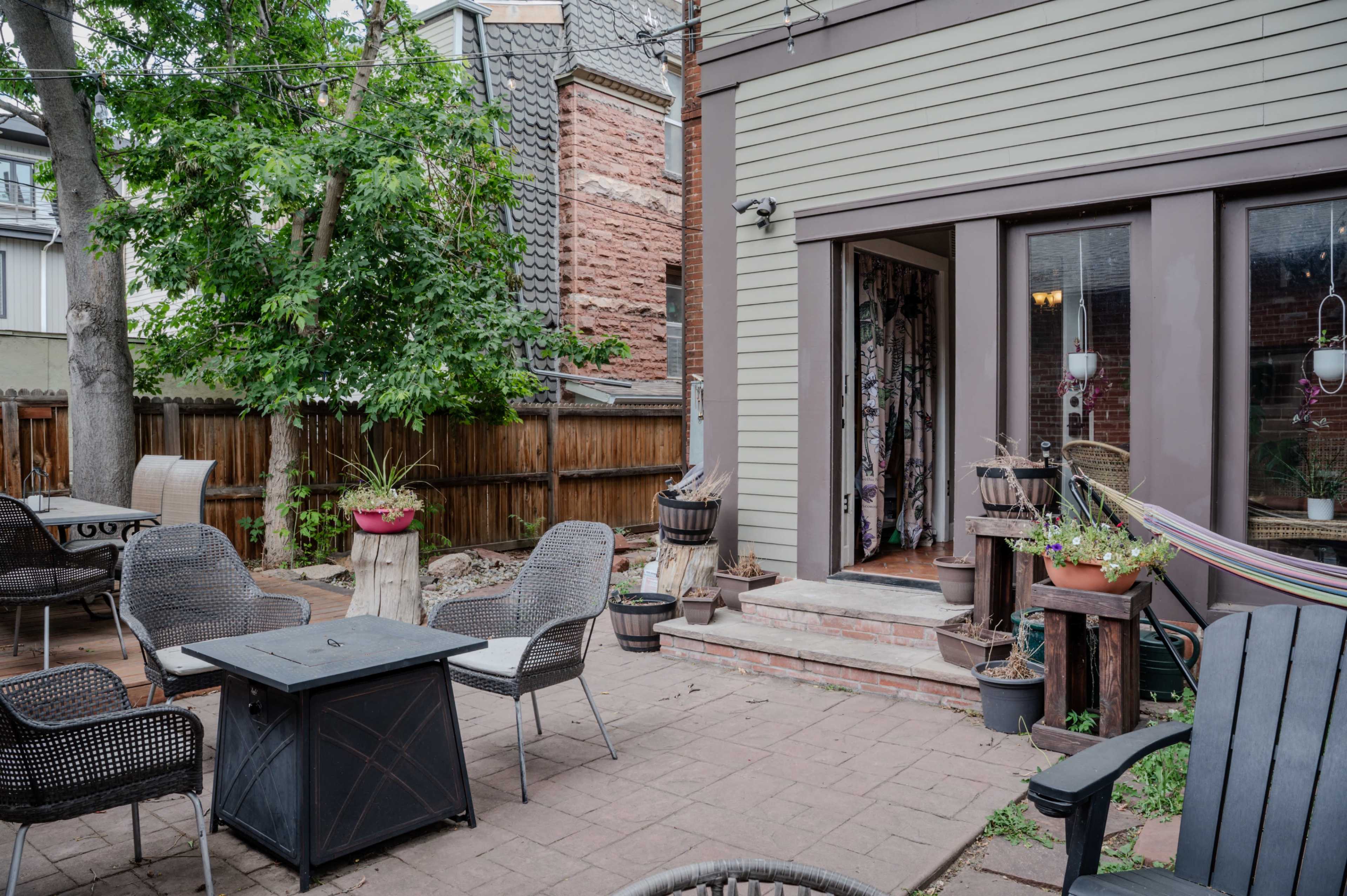 The image shows a patio area with outdoor furniture, including chairs and a table, surrounded by greenery and a wooden fence.