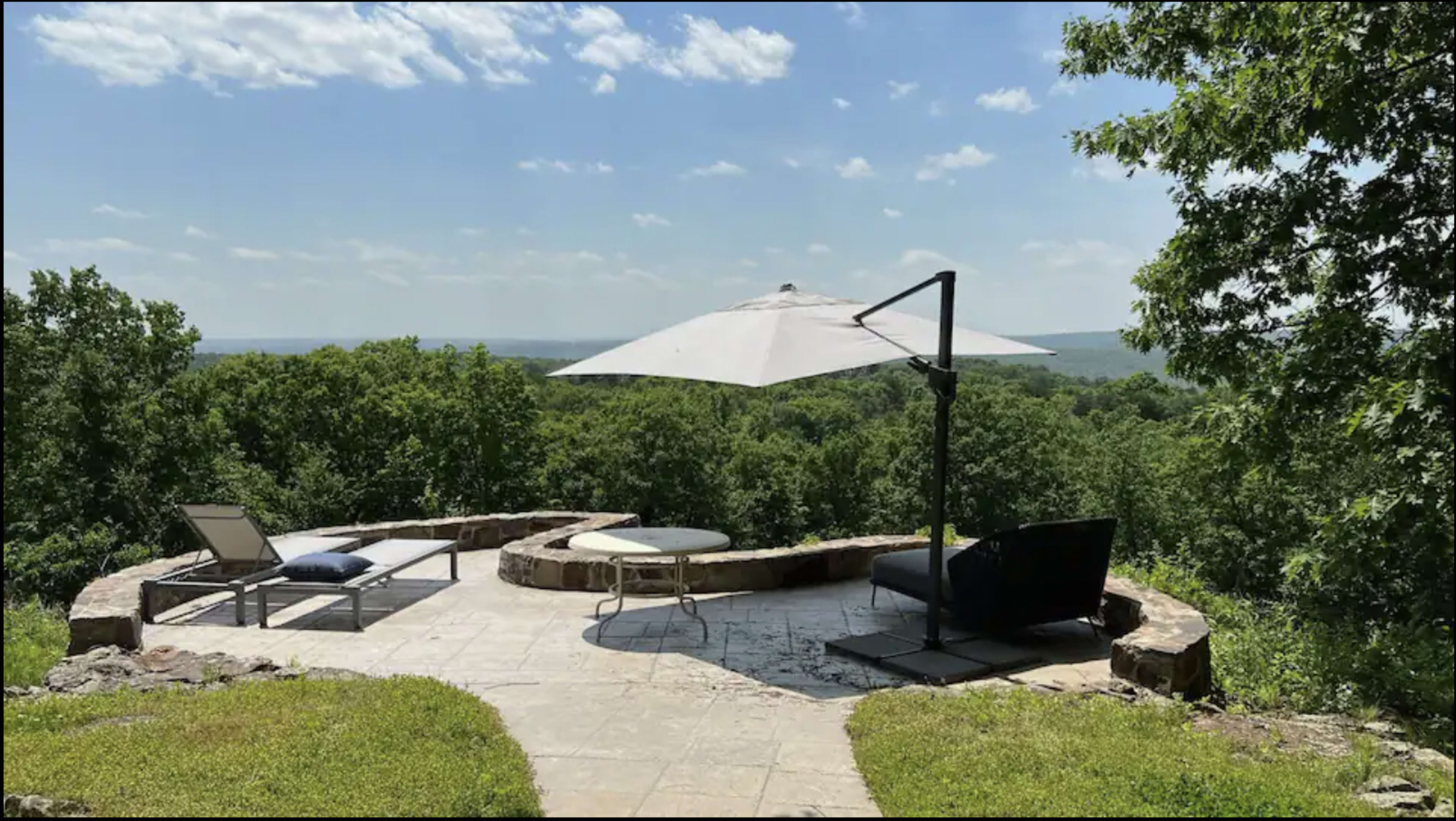 A stone patio overlooks a lush green landscape with a large umbrella and seating areas.