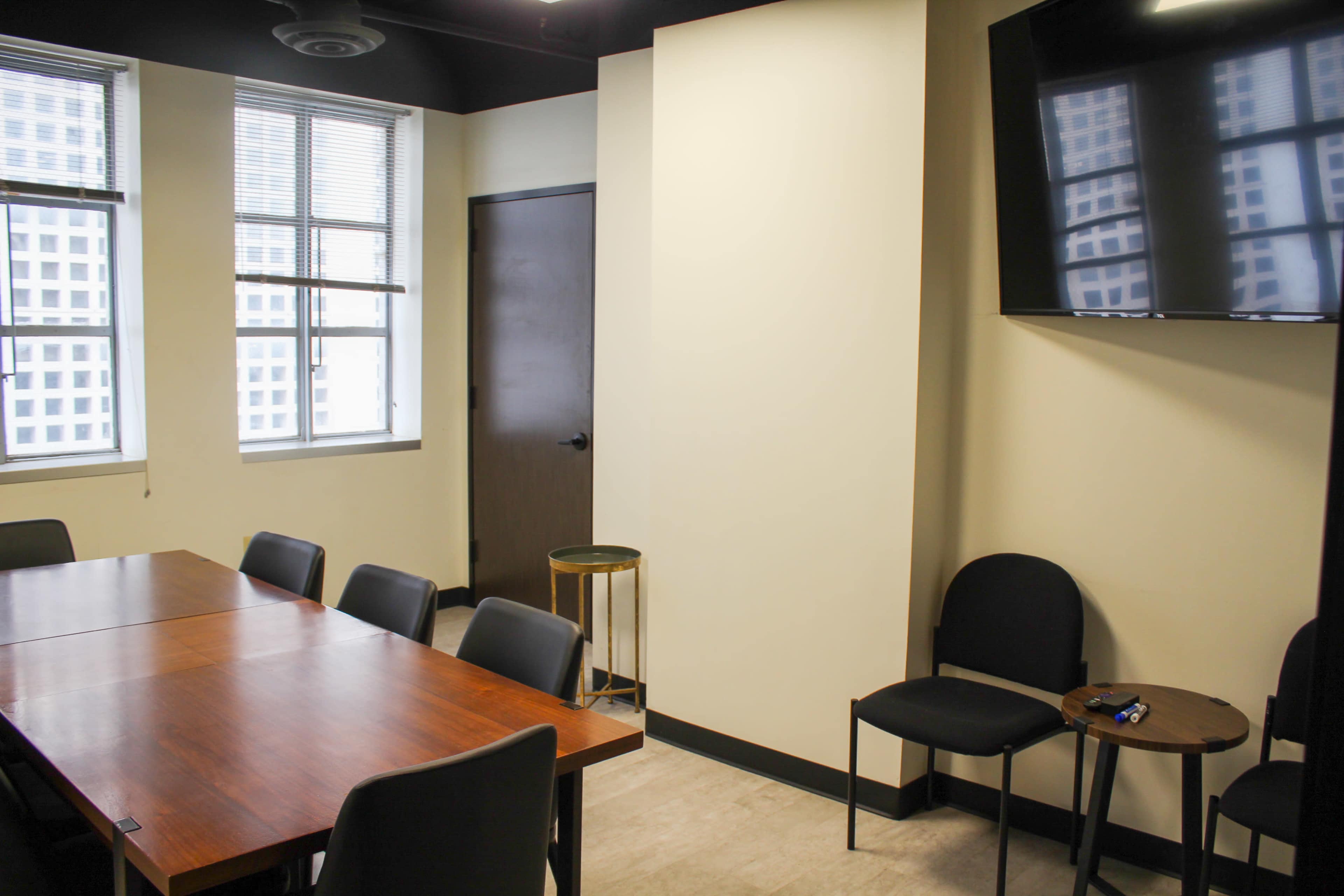 A conference room with a wooden table, several black chairs, a TV mounted on the wall, and large windows allowing natural light.