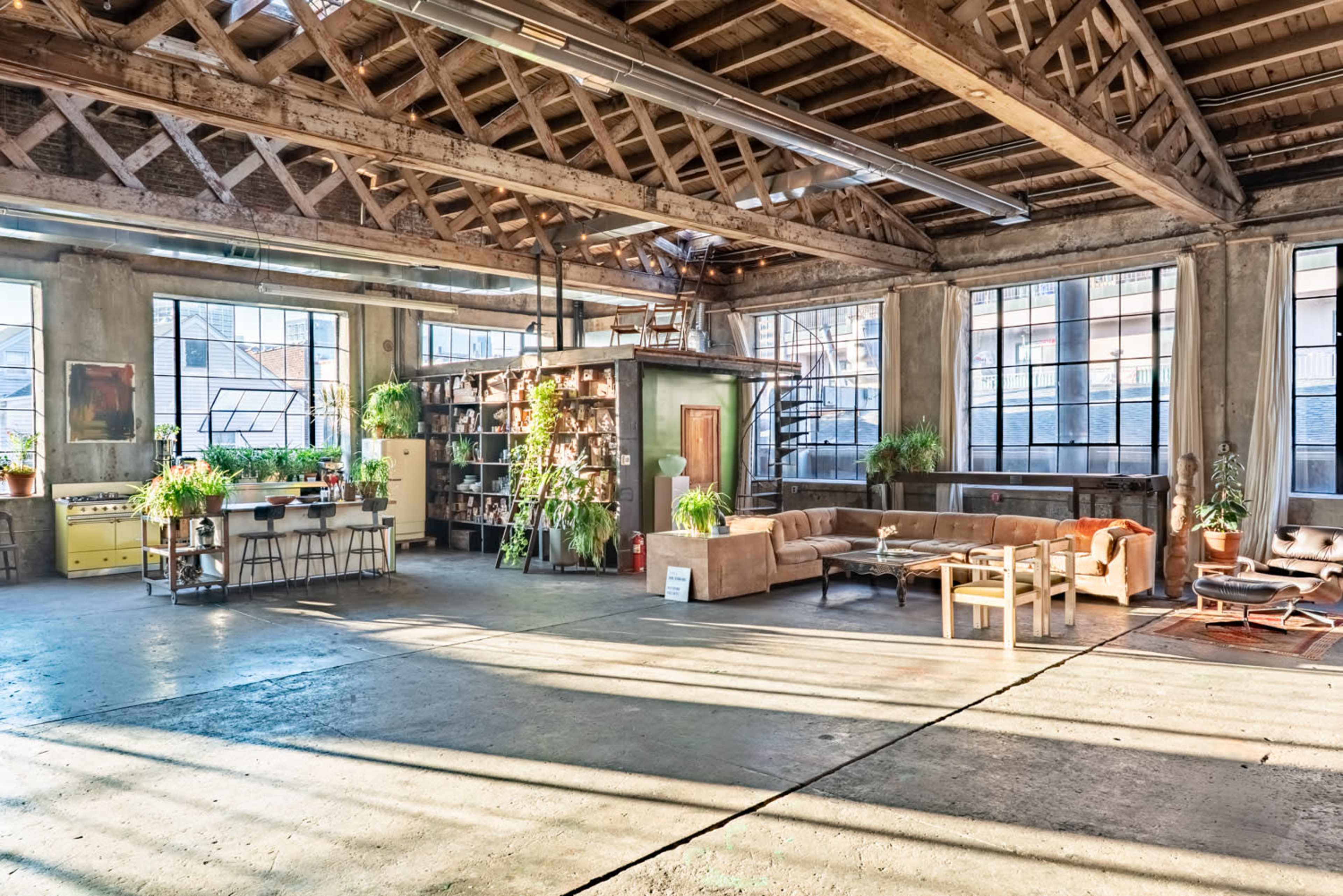 The image shows a spacious industrial-style loft with exposed wooden beams, large windows, and a mix of furniture surrounded by various plants.