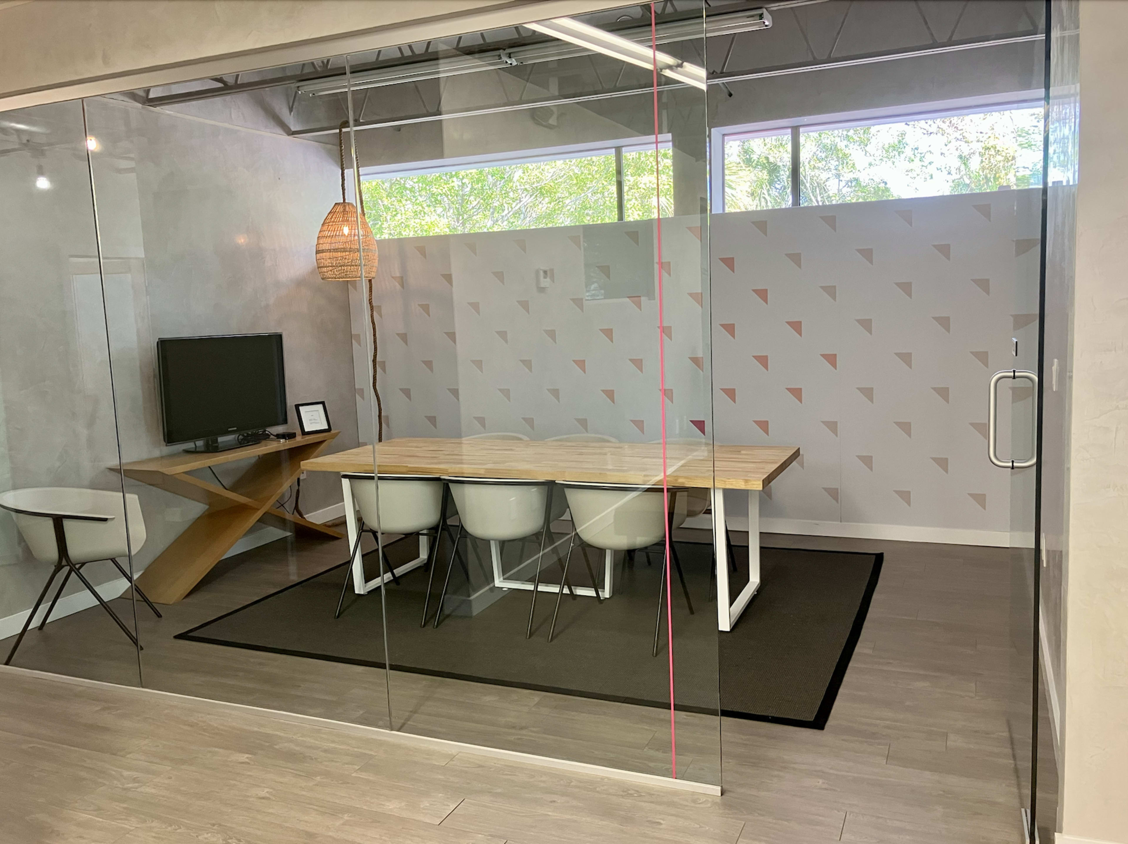 A modern conference room features a wooden table surrounded by white chairs, a television on the wall, and large glass windows with natural light.