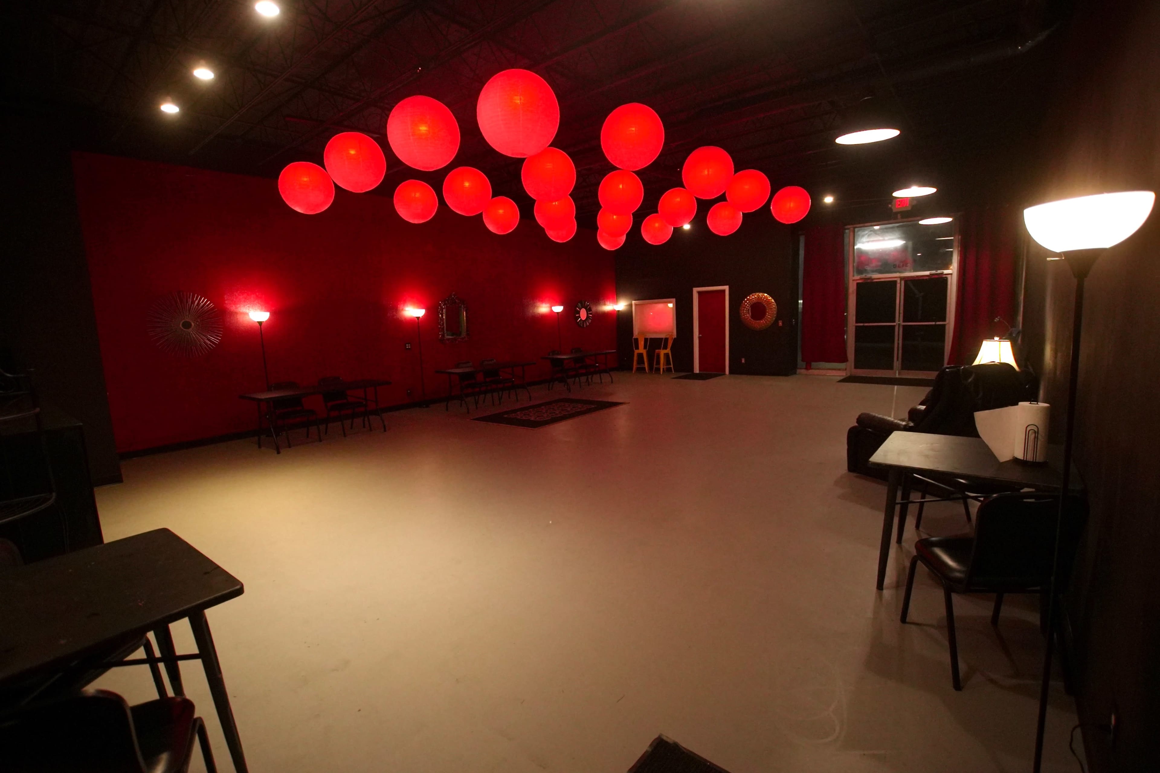 A dimly lit room with red walls, round red light fixtures hanging from the ceiling, and several tables and chairs arranged throughout the space.