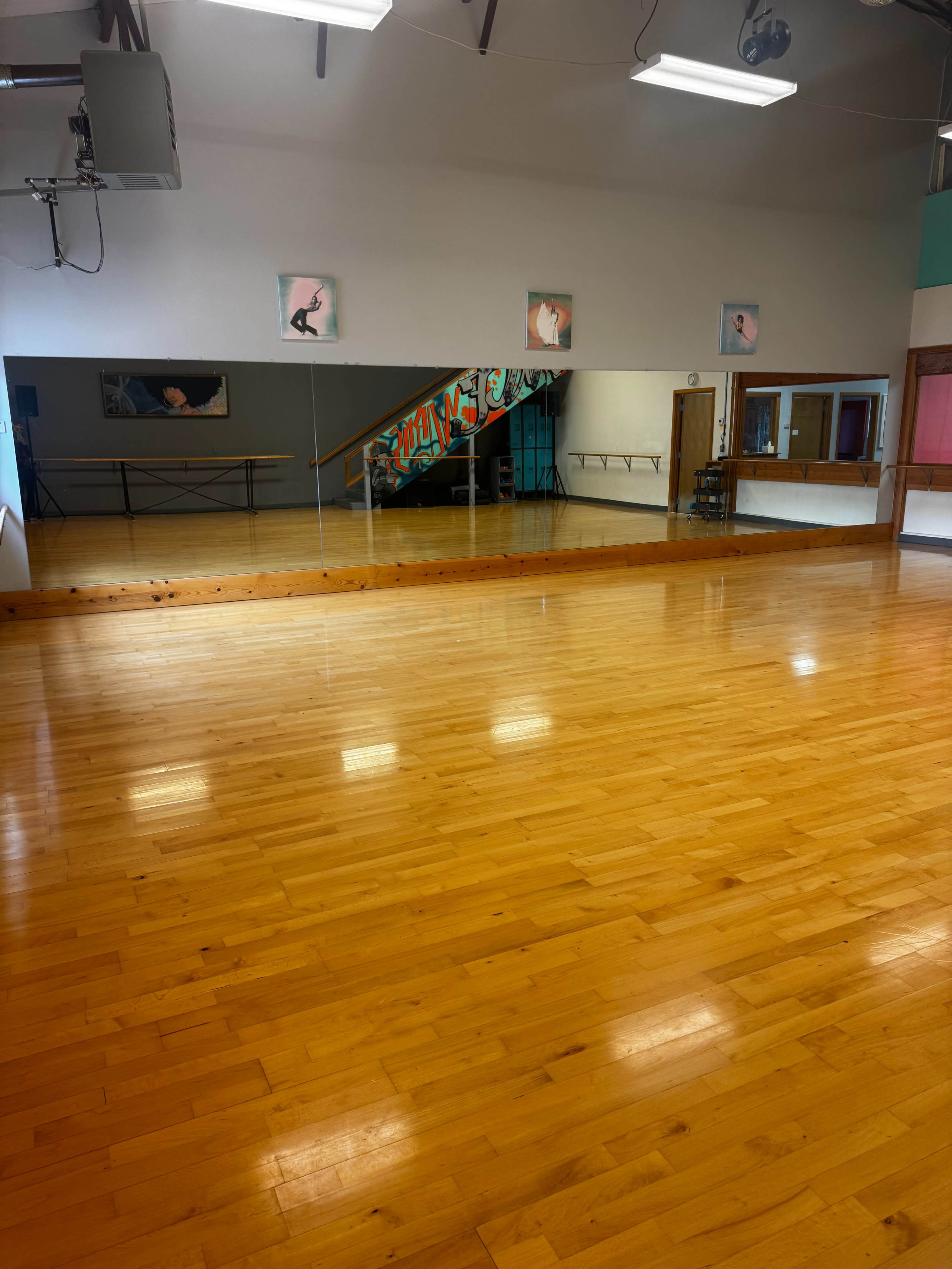 The image shows a spacious dance studio with a polished wooden floor, mirrors on one wall, and colorful artwork on the opposite wall.