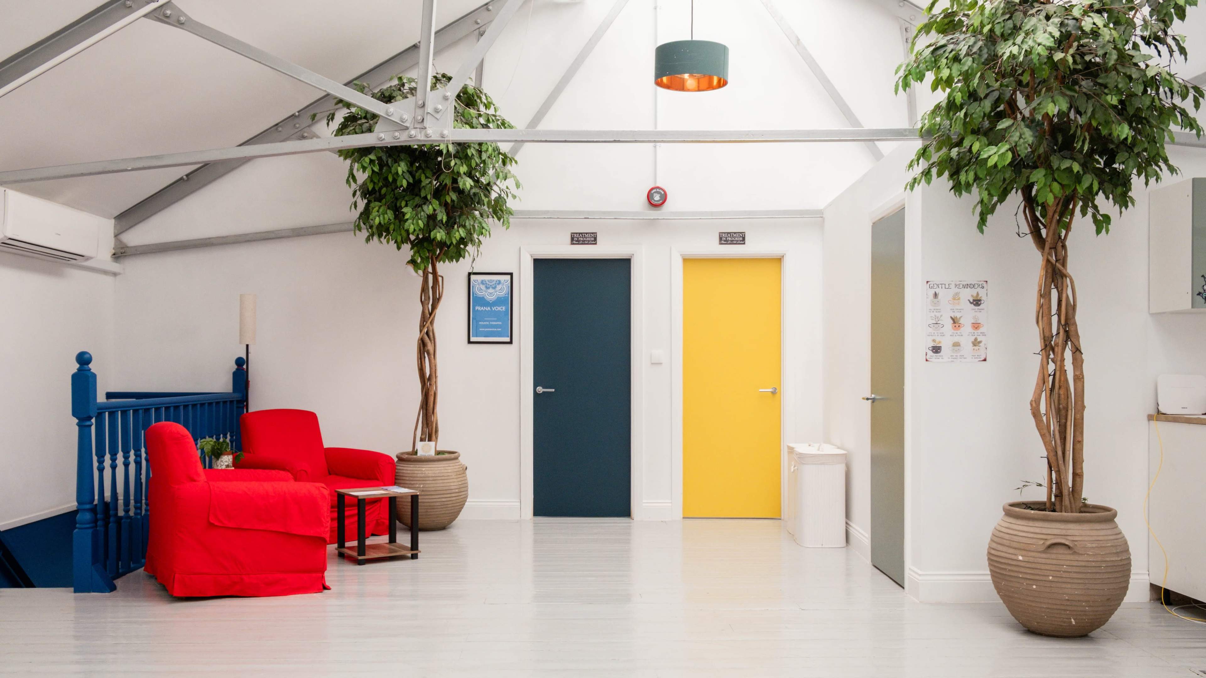 The image shows a bright, open interior space featuring a red sofa, two large potted trees, and colorful doors: one yellow and one gray.