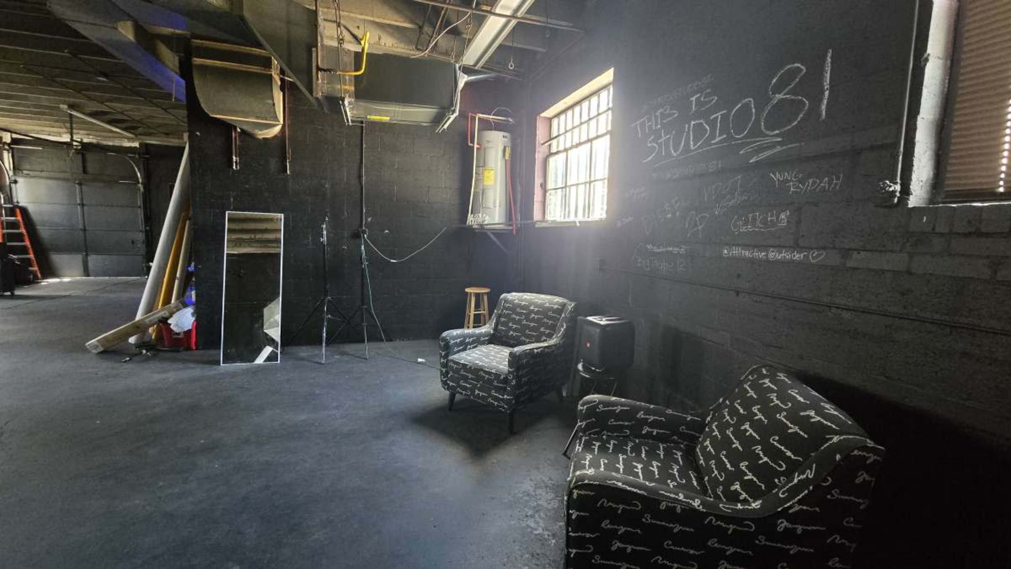 The image shows a dimly lit studio space with two patterned chairs, a mirror, and equipment, along with chalkboard-style writing on the wall.