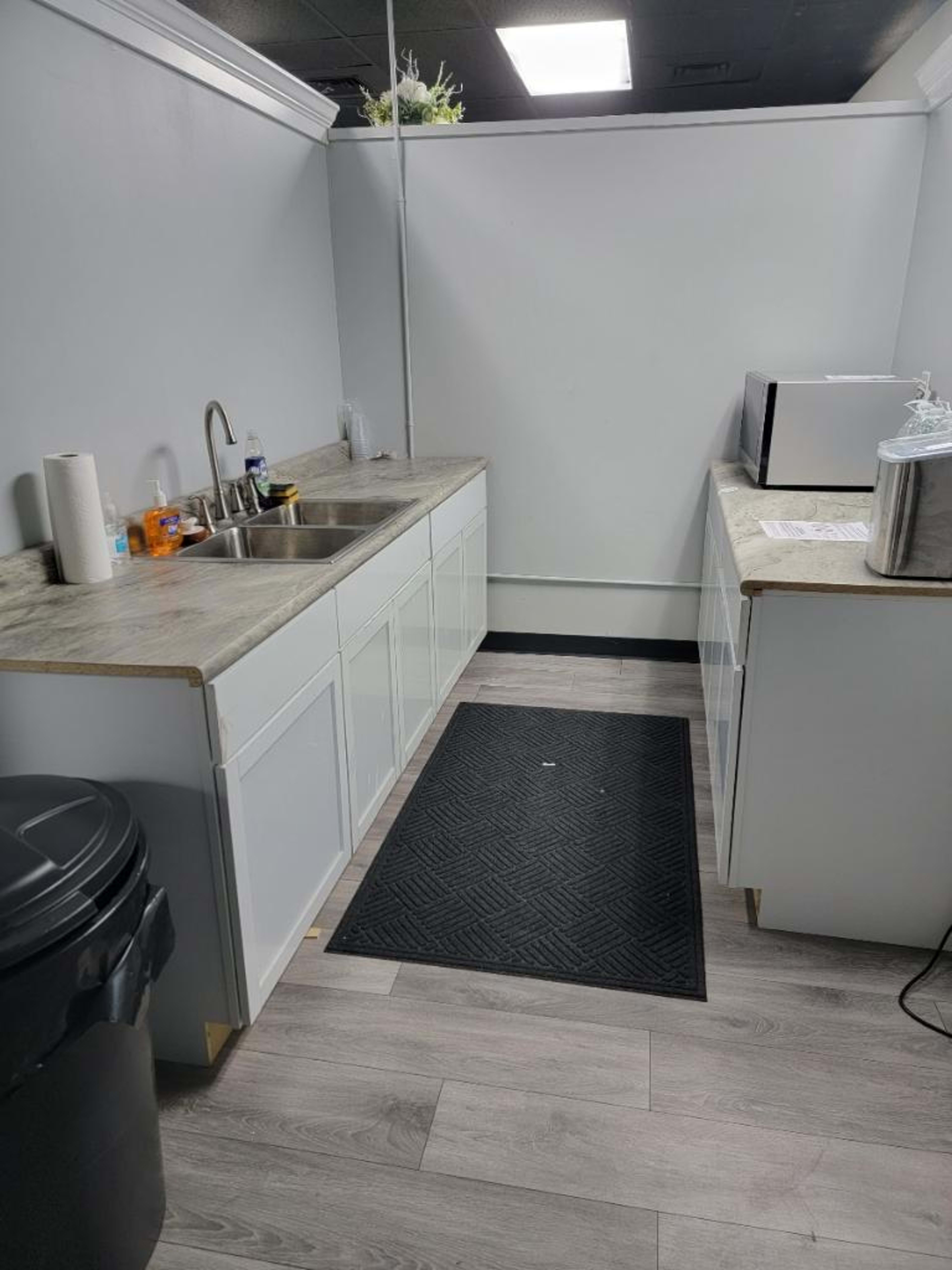 A small kitchen area featuring a sink, countertop, microwave, and a black trash can against a gray wall with light flooring.
