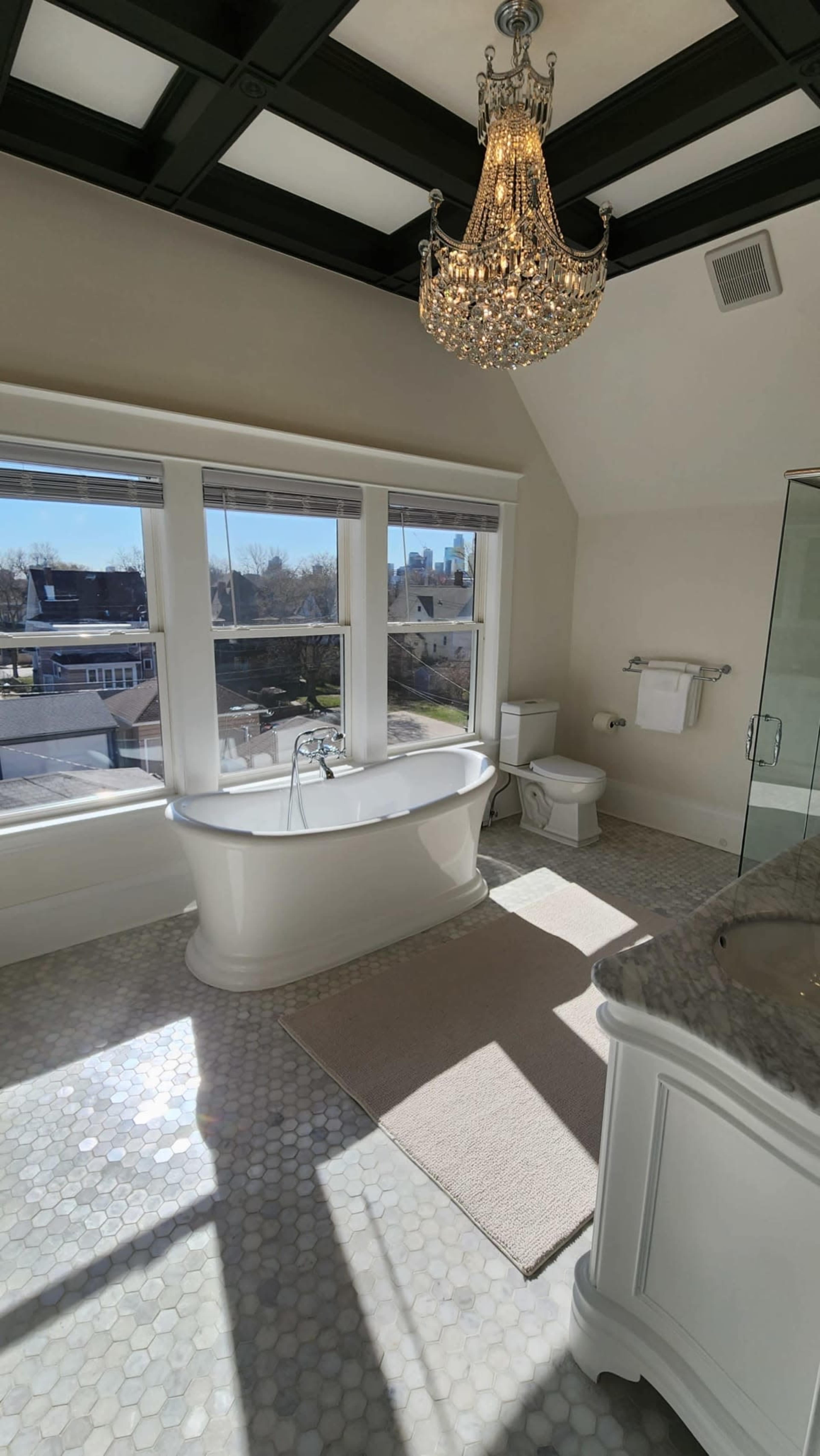 A bright bathroom features a freestanding bathtub, a chandelier, and large windows with a city view, complemented by hexagonal tile flooring.