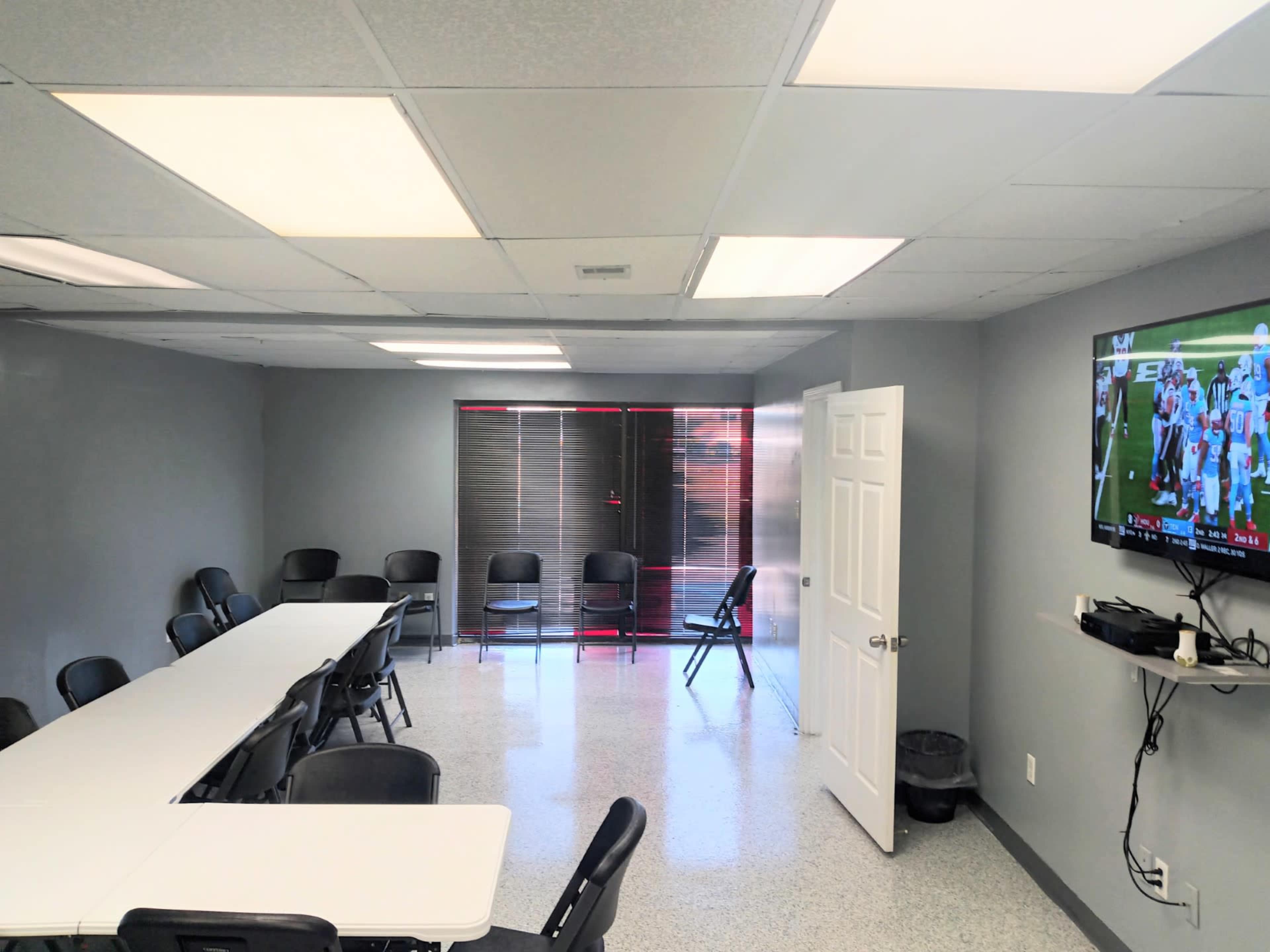 A meeting room with a long white table, black chairs, and a television mounted on the wall displaying a sports game.