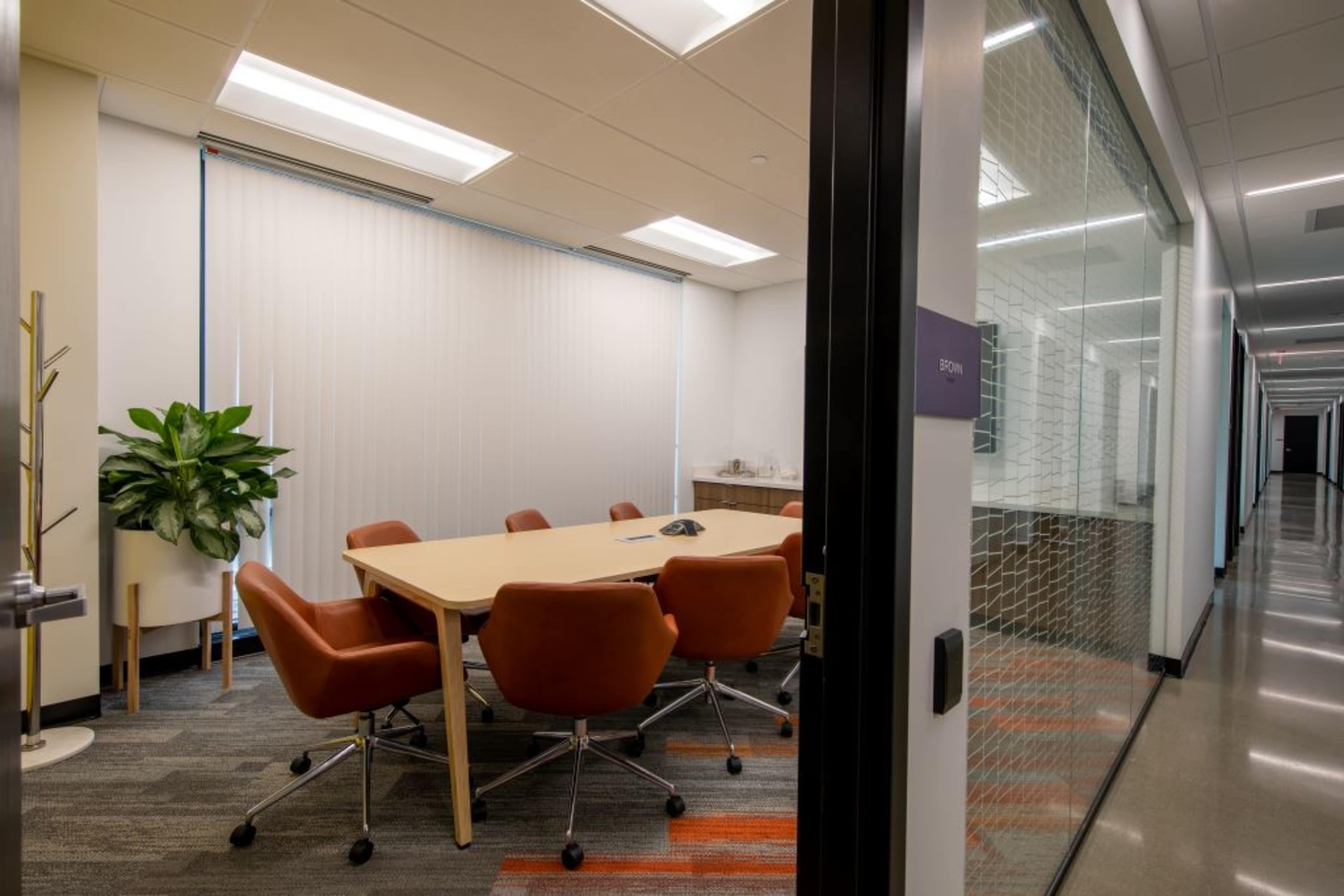 A conference room with a round table and six orange chairs, featuring a large plant and a phone on the table, is visible through a glass door.