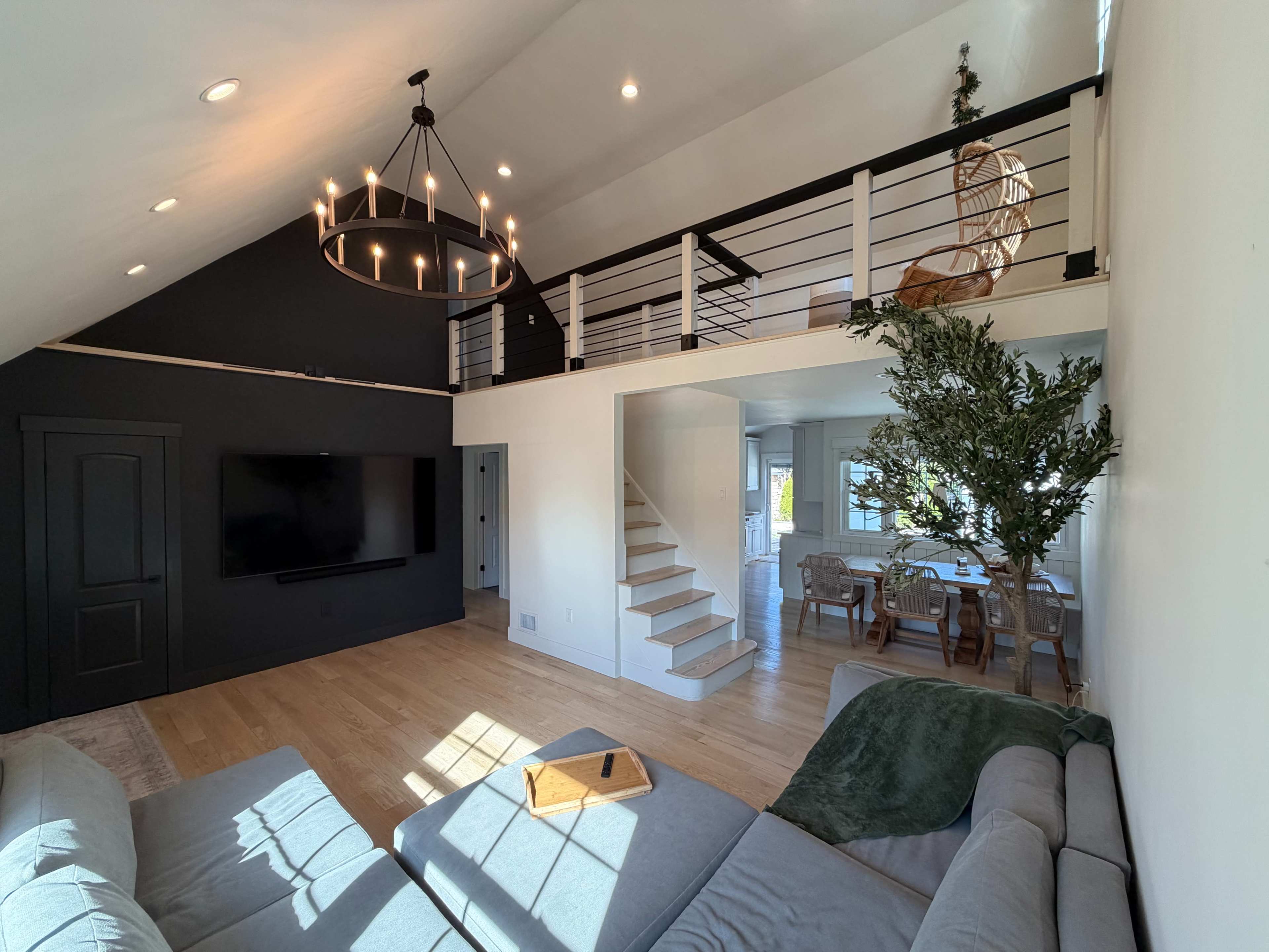 The image depicts a modern living room with a gray sectional sofa, a large television mounted on a black wall, and a staircase leading to an upper loft area.