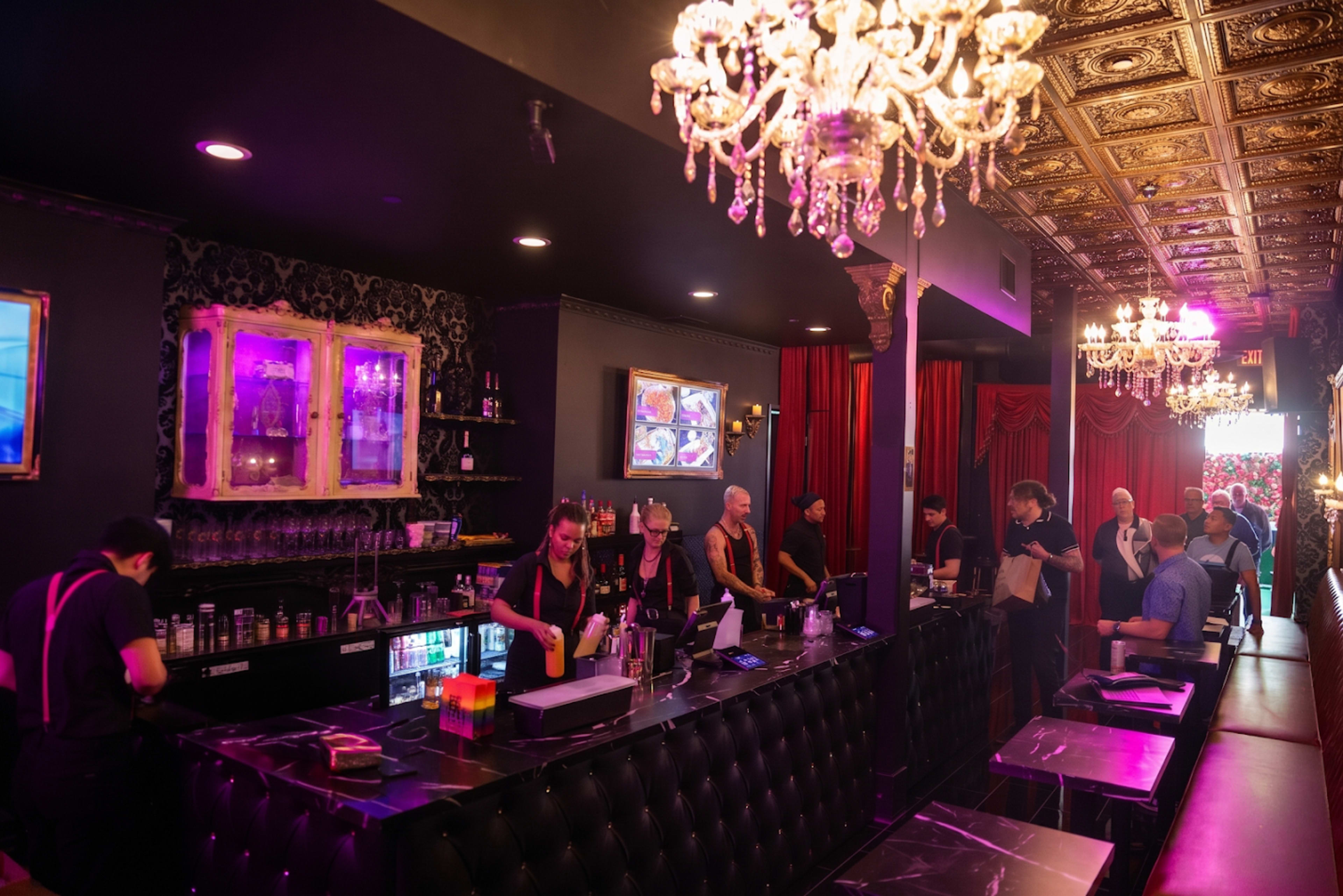 A dimly lit bar features a long black counter with staff preparing drinks, surrounded by ornate chandeliers and red drapery.