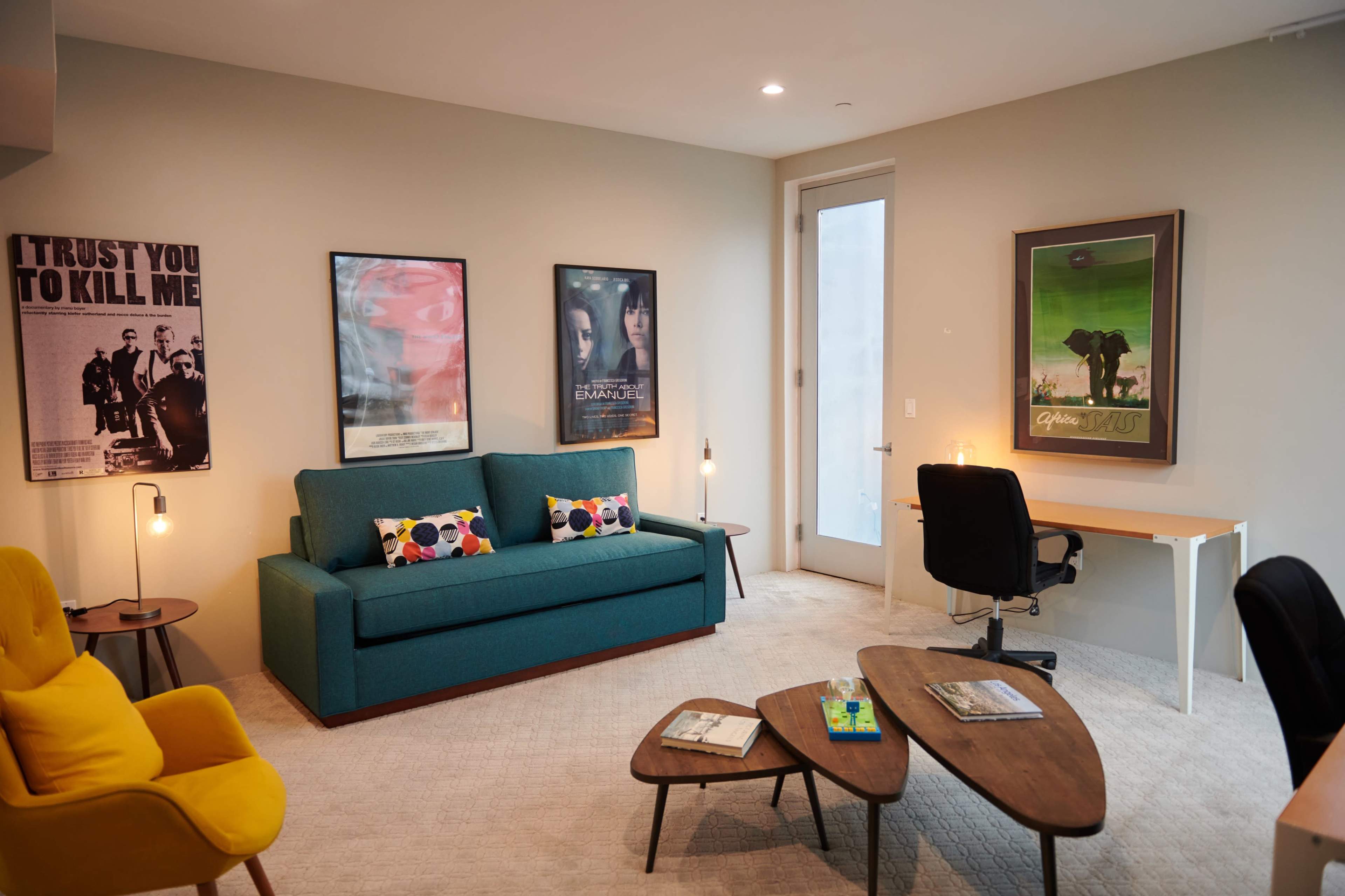 A modern living room with a turquoise sofa, wooden coffee tables, decorative artwork on the walls, and a desk with a chair near a large window.