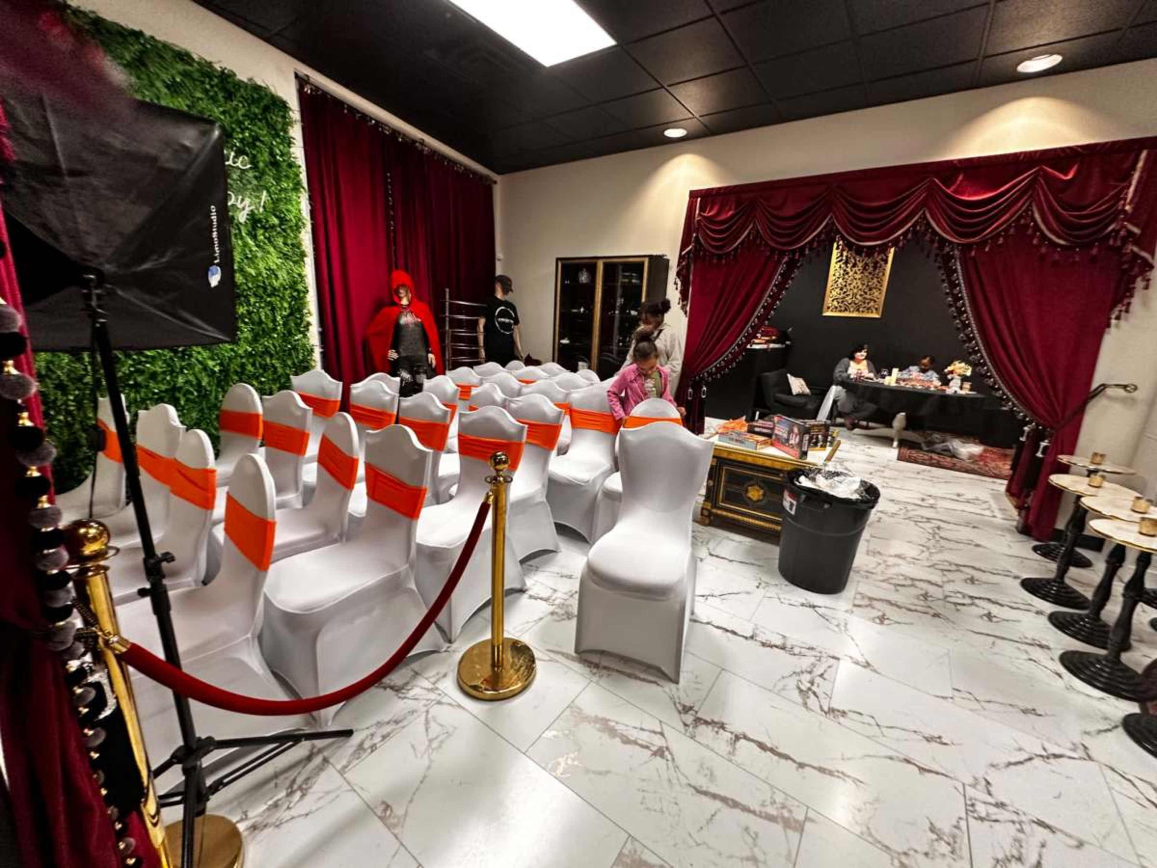The image shows a room set up for an event with white chairs adorned with orange ribbons, a red curtain backdrop, and a small table in the corner with people seated at it.