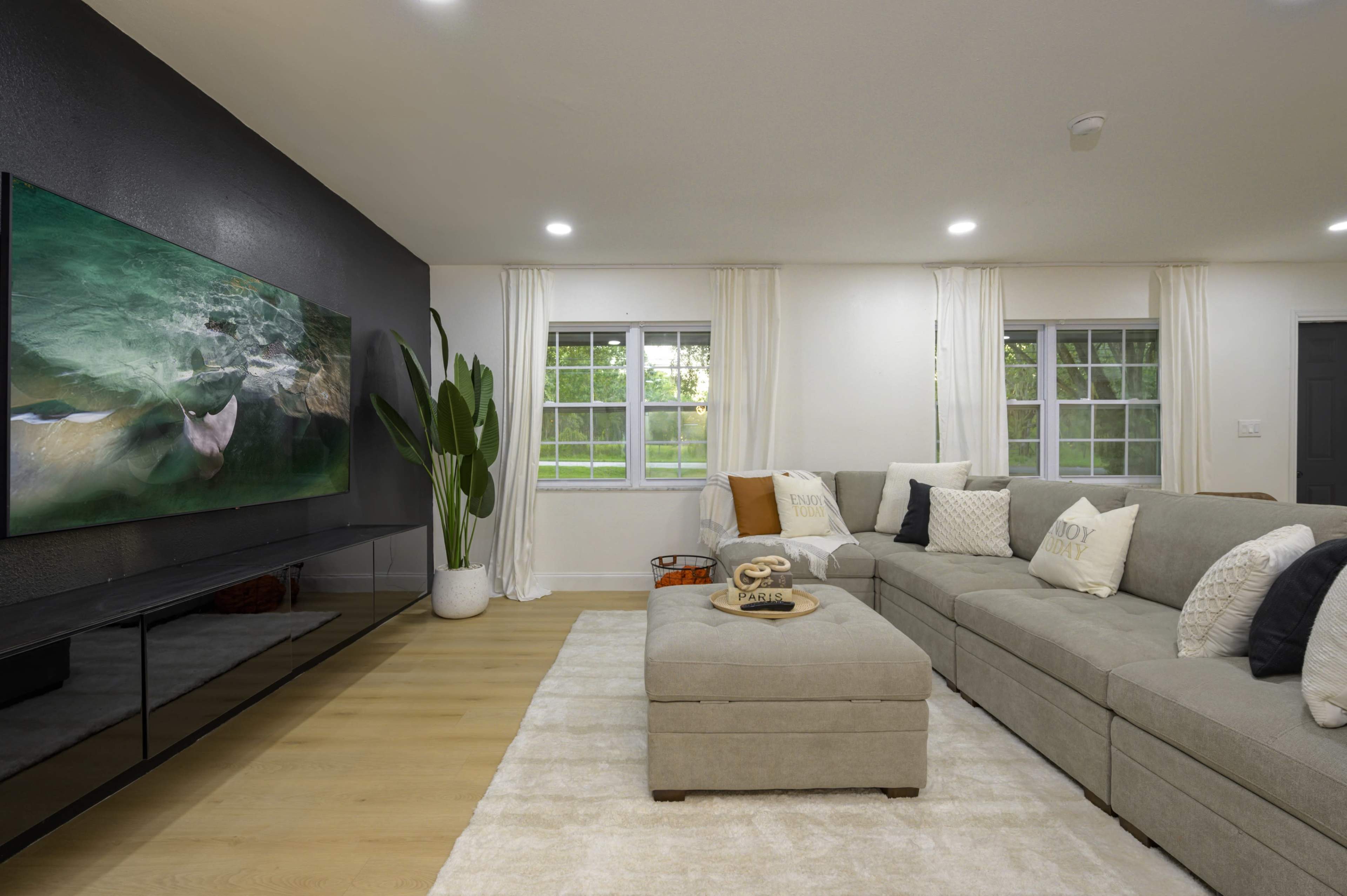 A modern living room with a large sectional sofa, a flat-screen TV mounted on a black accent wall, and large windows allowing natural light.