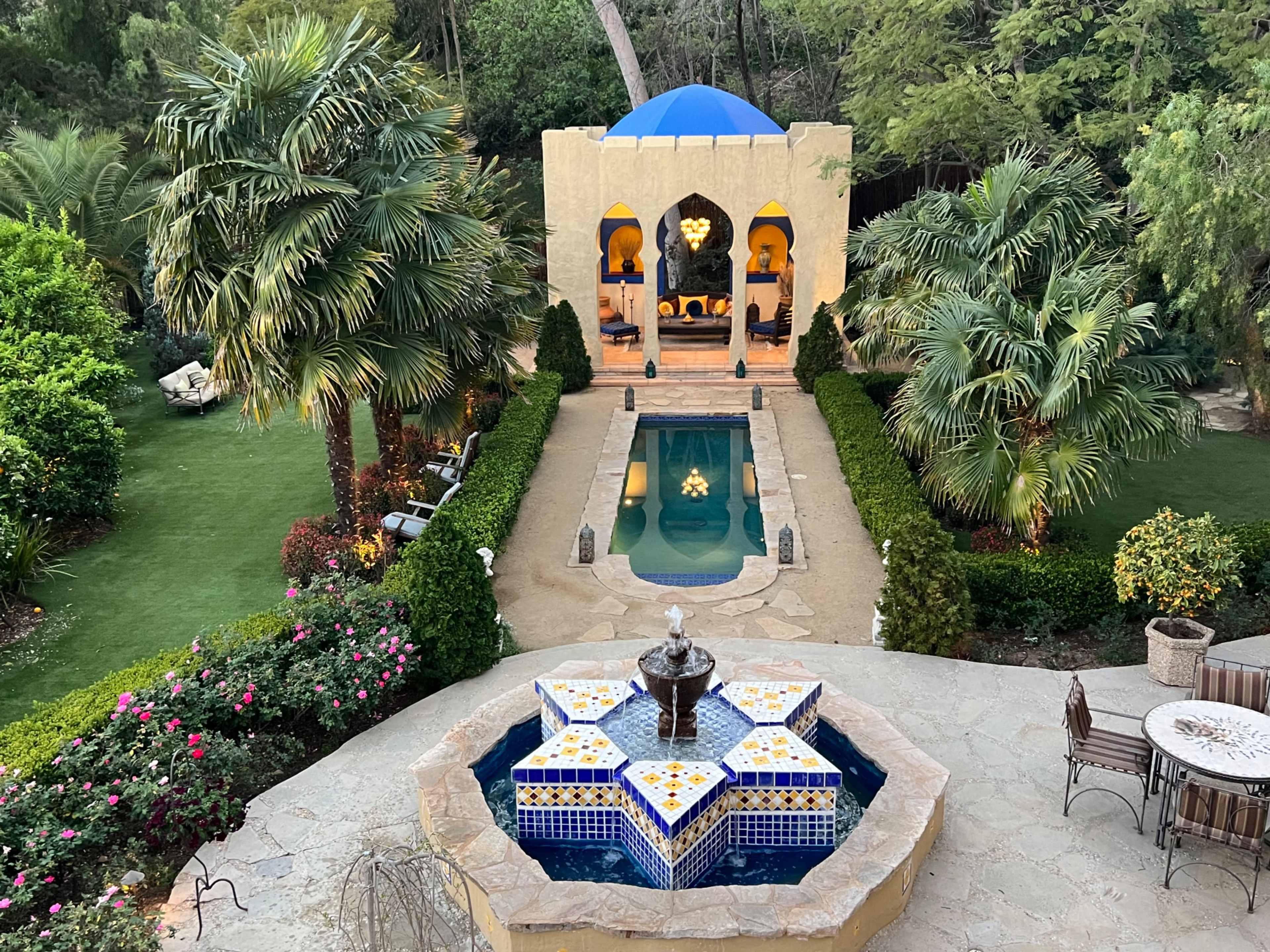 The image shows a landscaped garden featuring a swimming pool, an ornate fountain at the forefront, and a pavilion set amid palm trees and flowering plants.