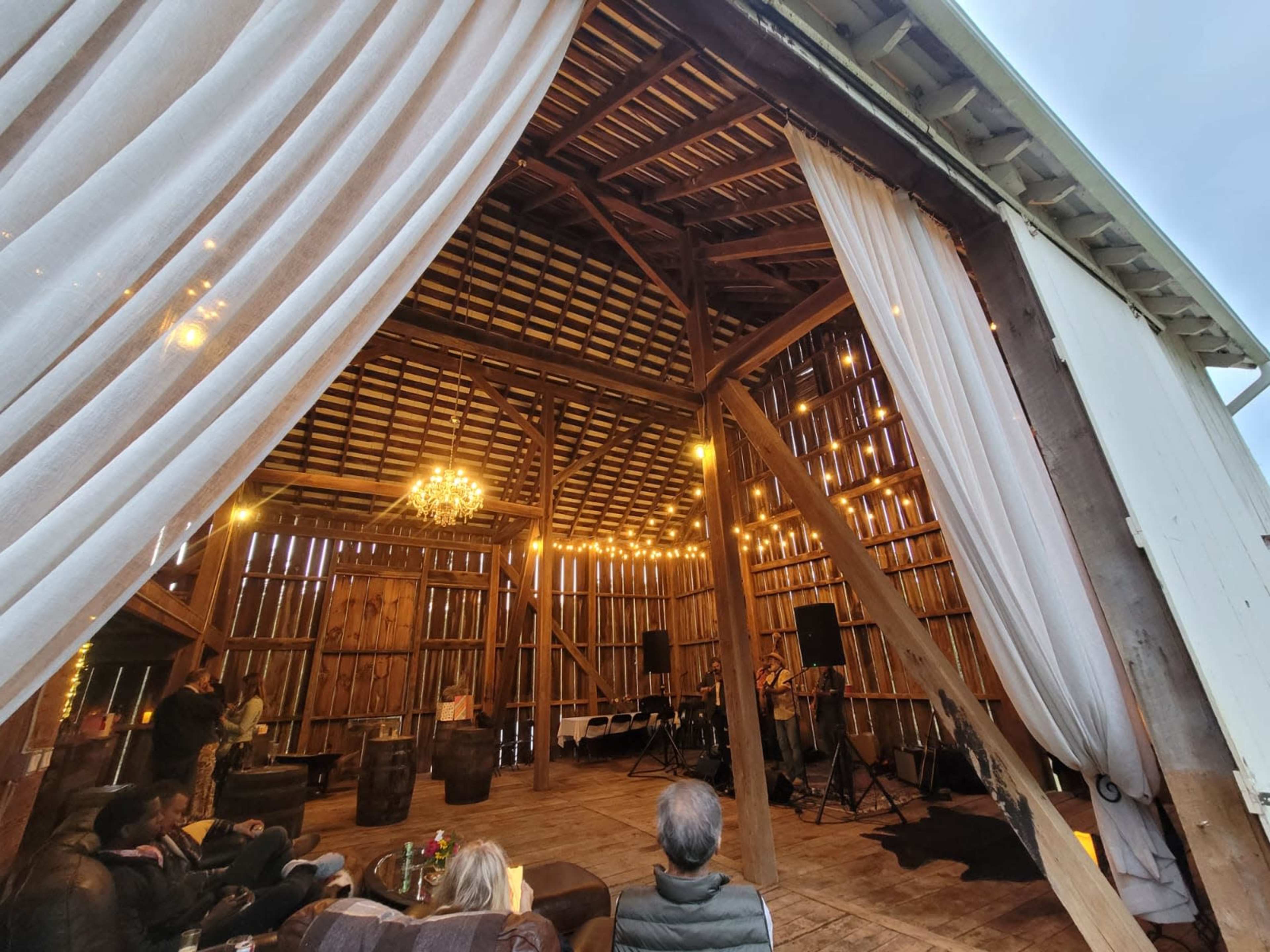 The interior of a large barn is decorated with draped curtains, string lights, and a chandelier, while guests sit on sofas and enjoy live music.
