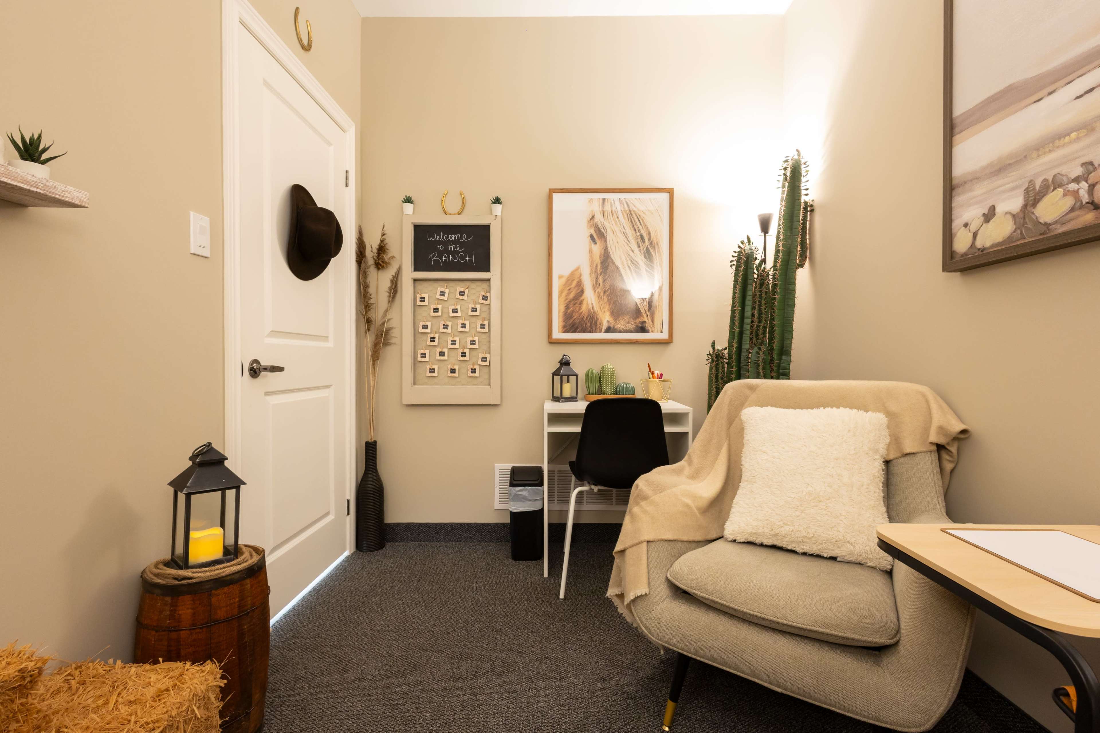 The image shows a small, decorated room with a chair, desk, wall art, and plants, featuring a door and a few accessories like a lantern and a hat.