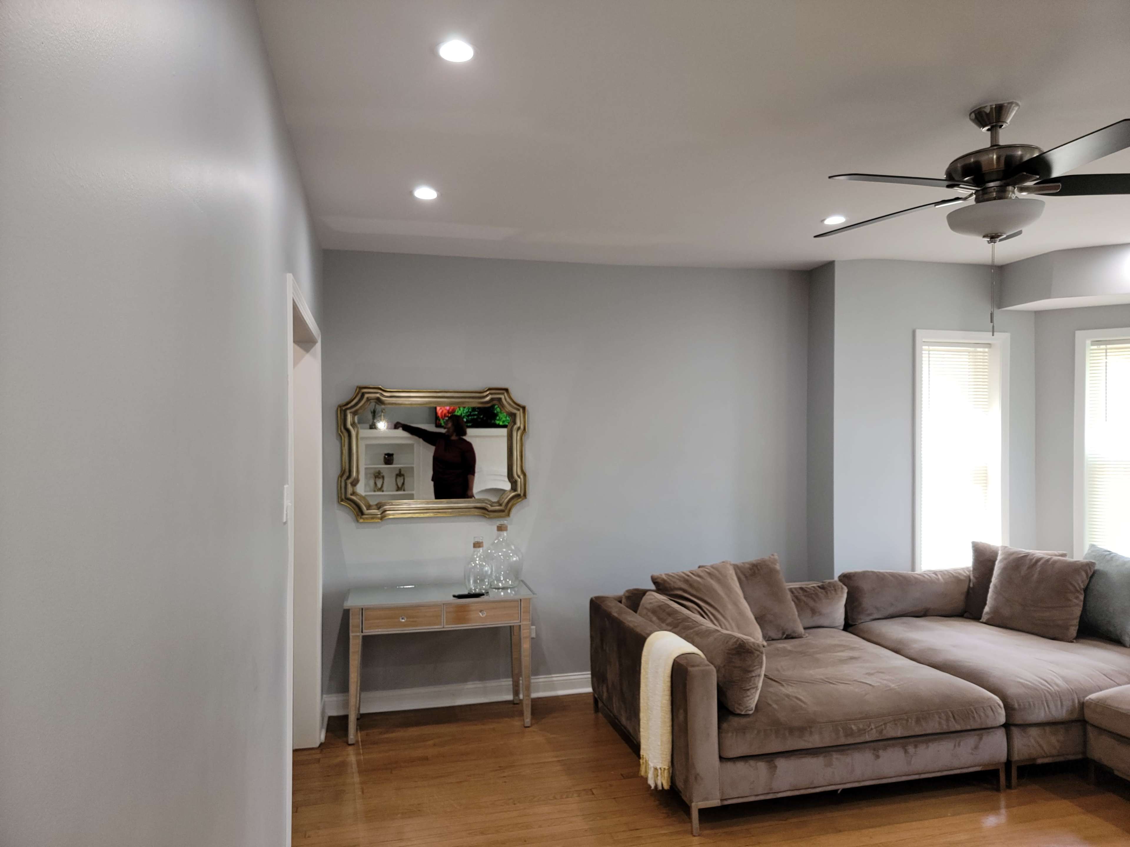A modern living room features a brown sectional sofa, a decorative mirror on the wall, and a small table with glass bottles.