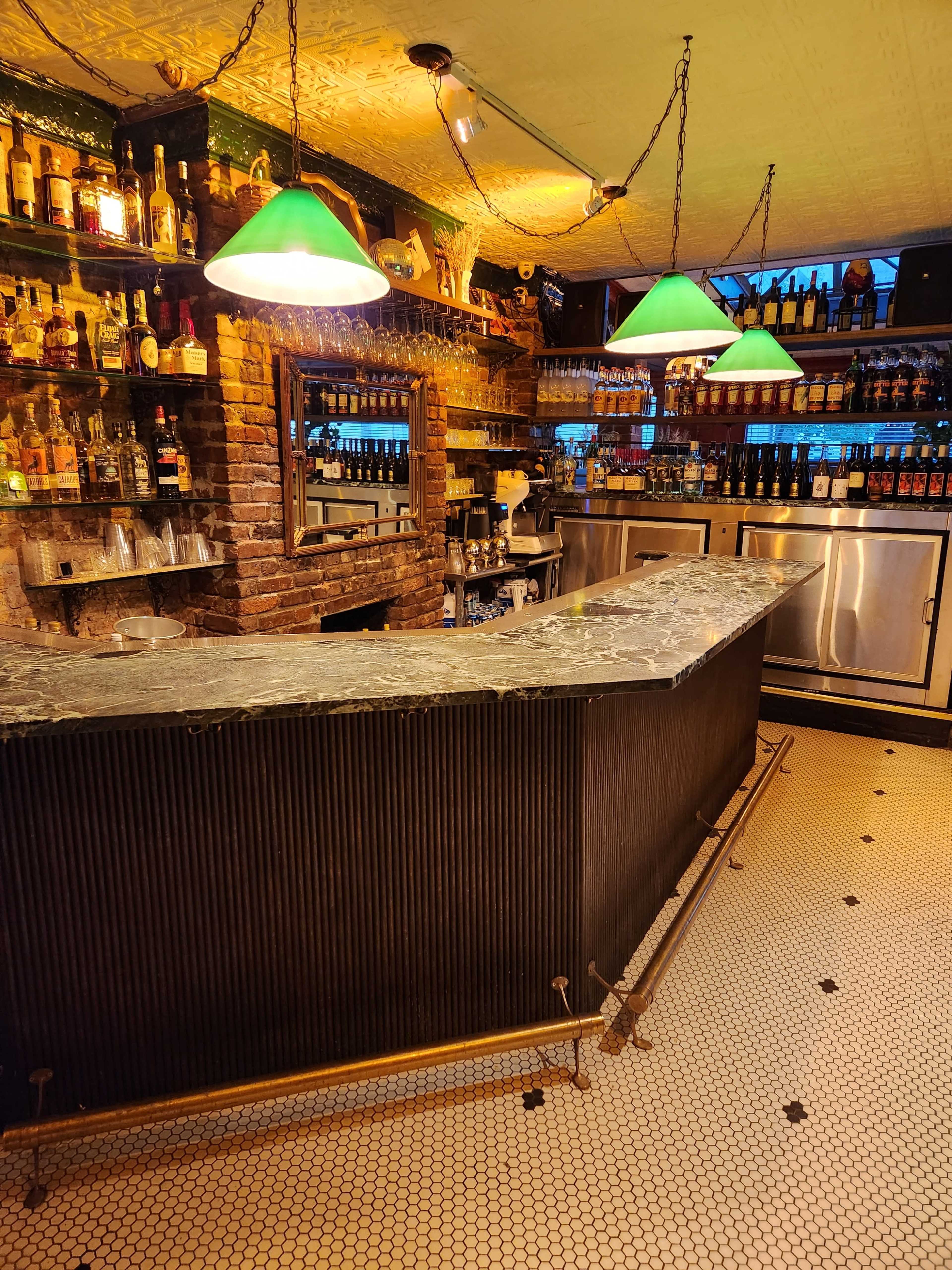 The image shows a bar with a dark stone countertop, green pendant lights, and exposed brick walls lined with various bottles and glasses.