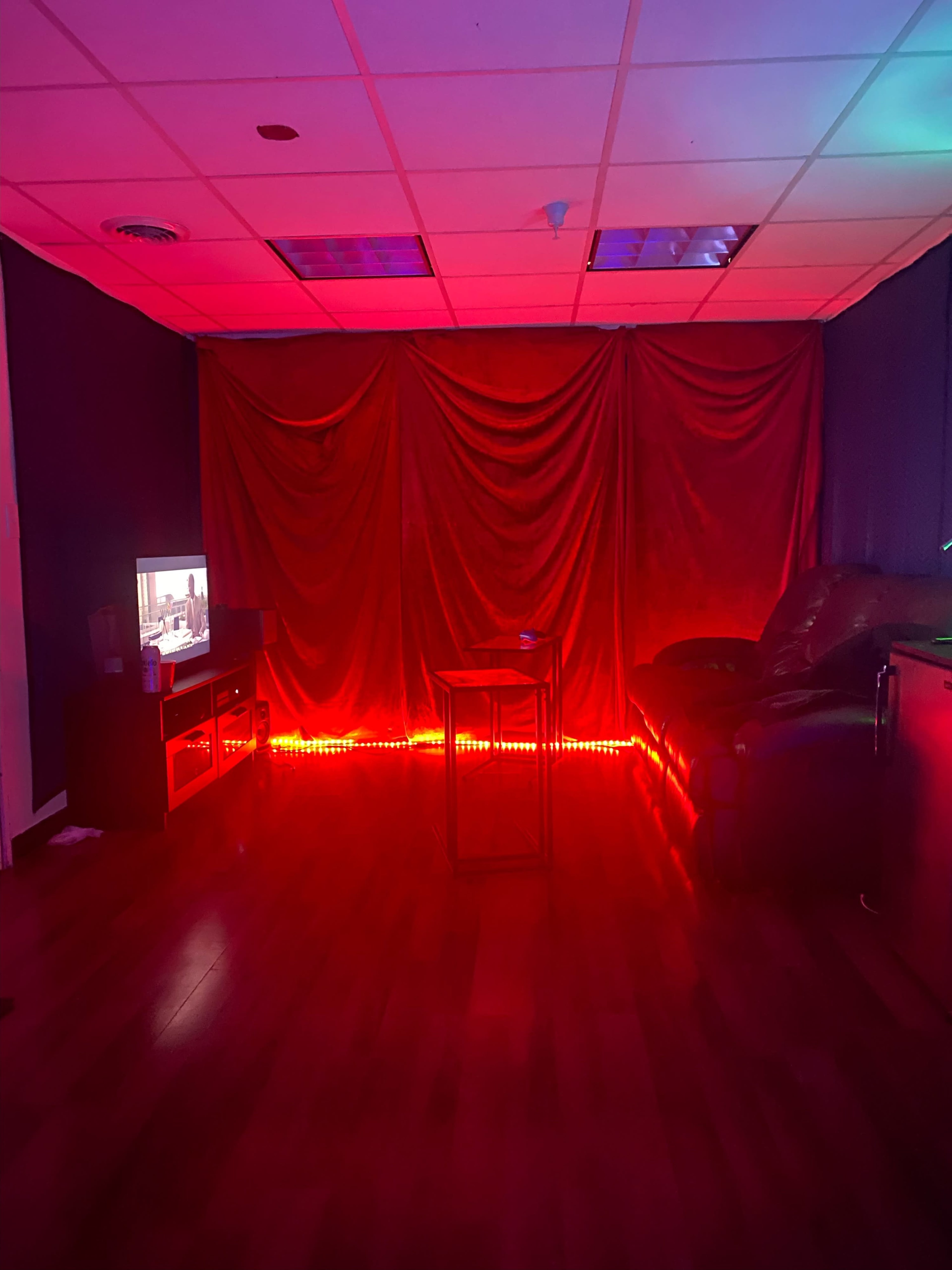 The room features a red-lit area with a couch, a small table, and a television against a backdrop of draped fabric.