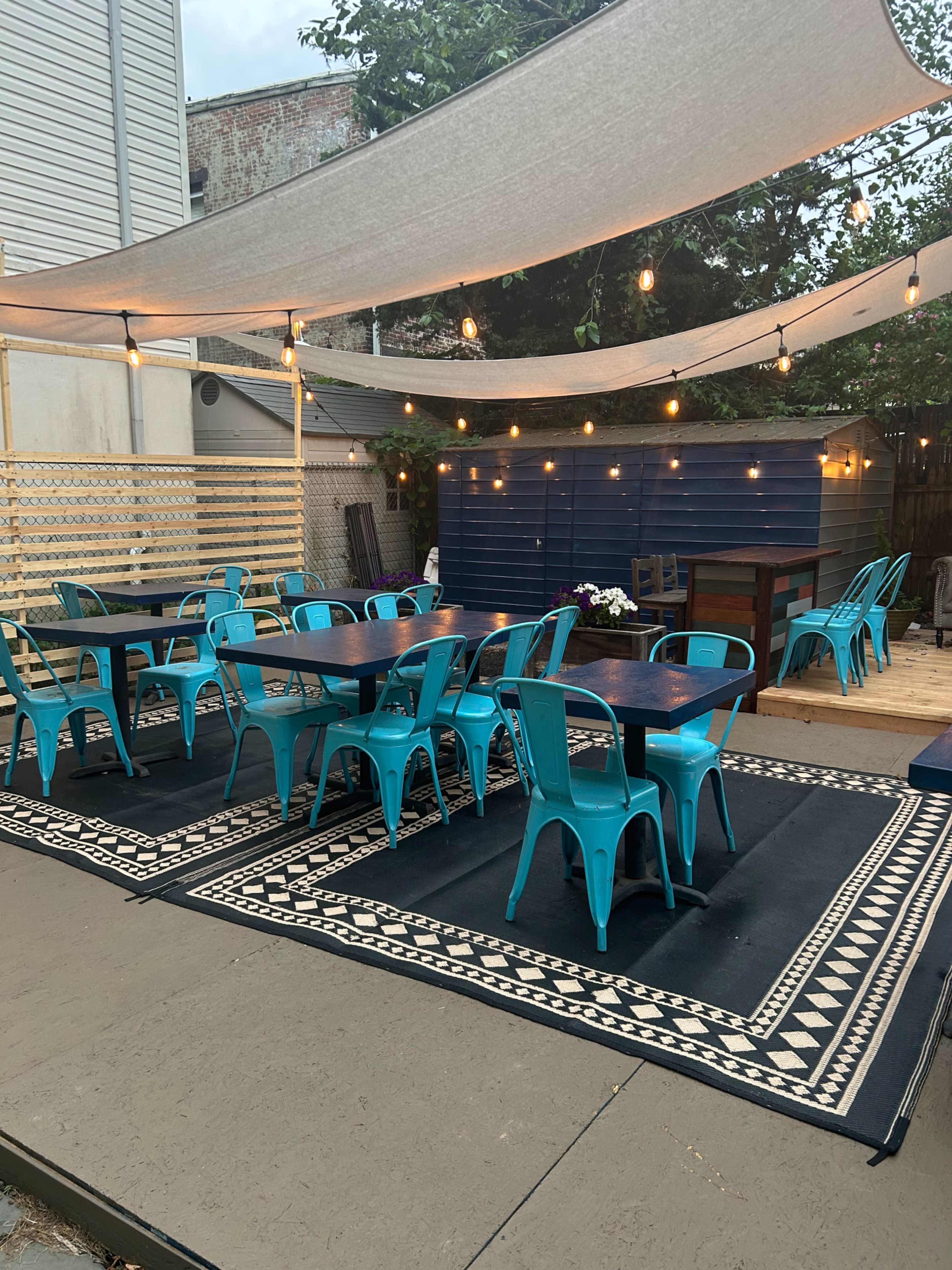 The outdoor space features several turquoise tables and chairs beneath a fabric canopy, with string lights overhead and patterned rugs on the ground.