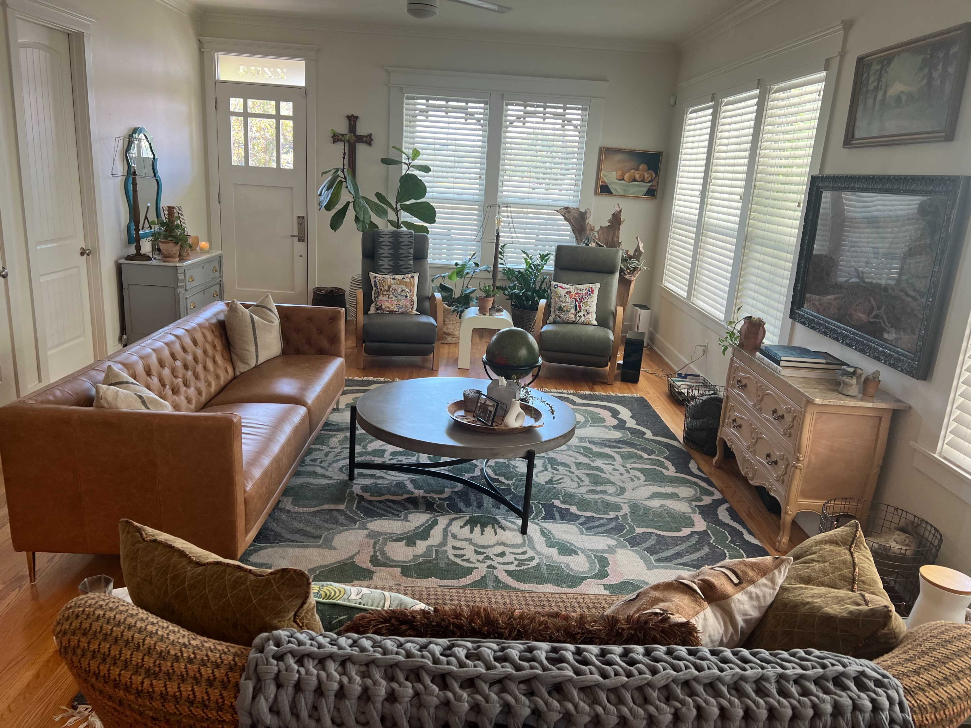 The living room features a leather couch, a circular coffee table, and two armchairs, all arranged around a patterned area rug with large windows allowing natural light to enter.