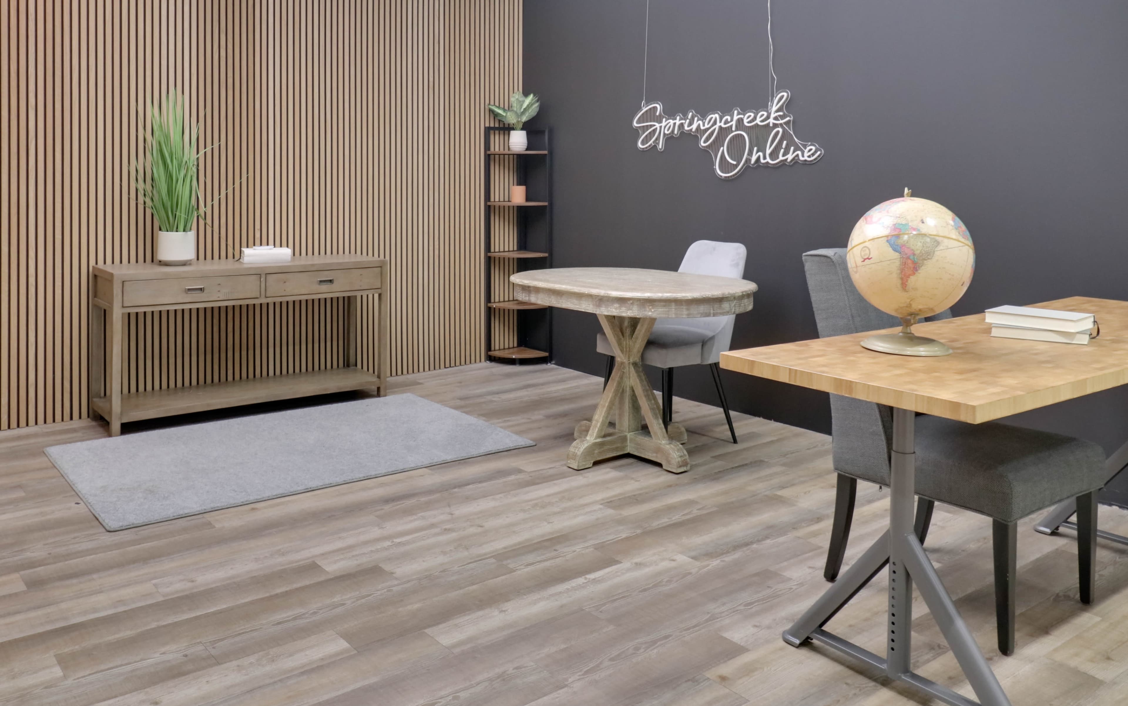 The image shows a minimalist office space featuring a wooden console table, a round table with a globe, a gray rug, and a plant on a shelf against a dark wall.