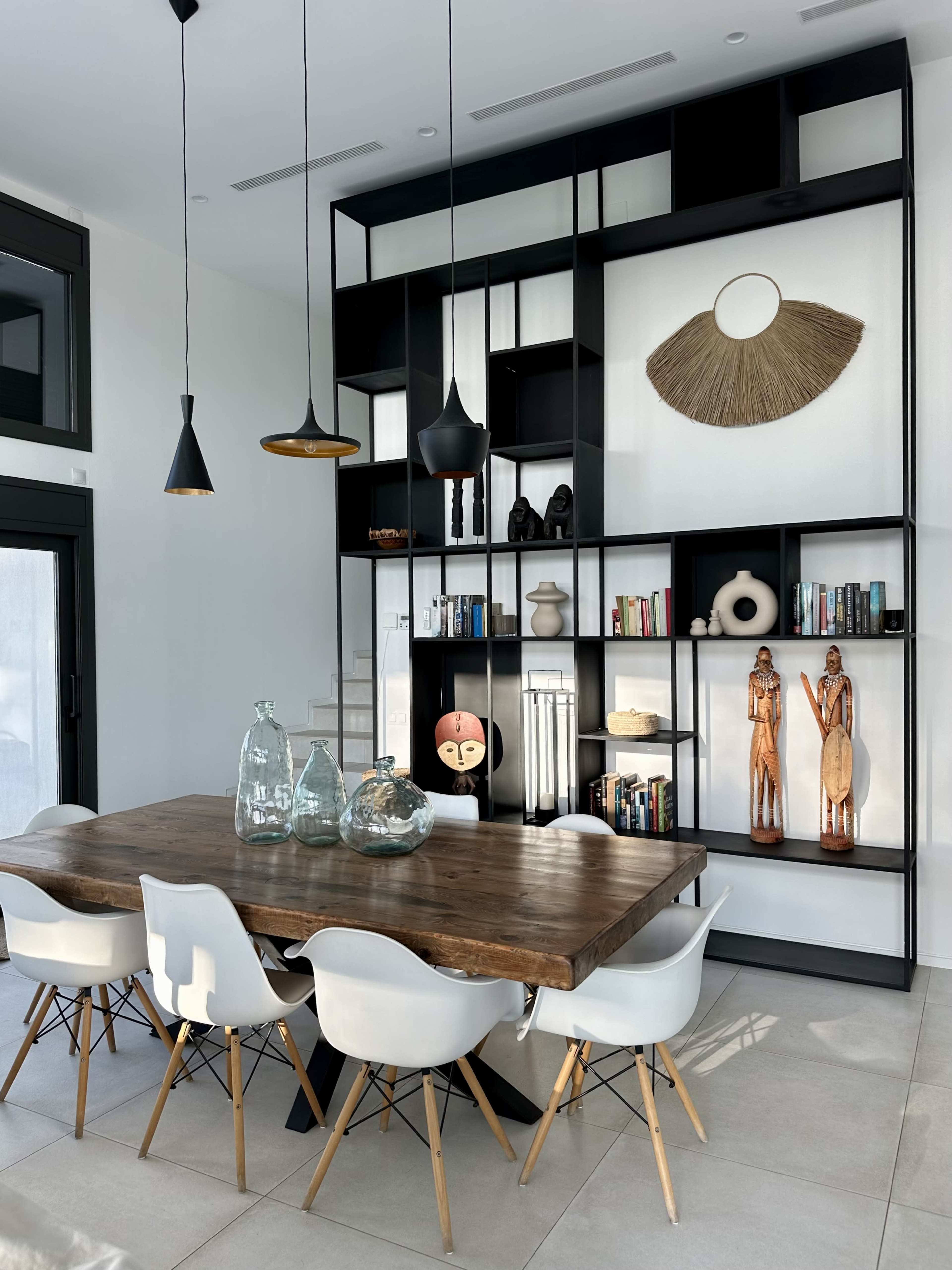 The image shows a modern dining area featuring a wooden table surrounded by white chairs, with a black bookshelf displaying decorative items and books in the background.