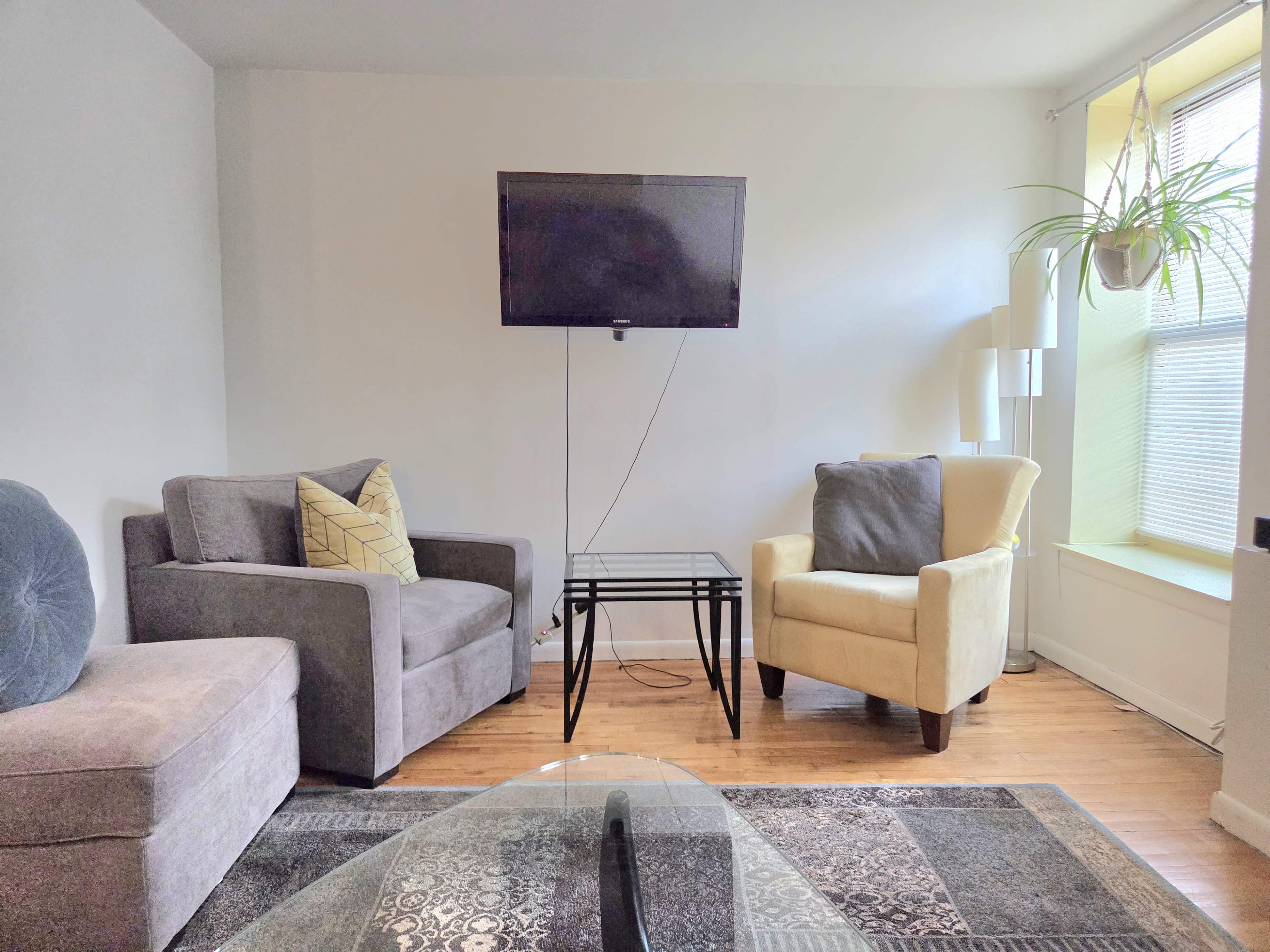 The image shows a small living room with two armchairs, a glass coffee table, and a wall-mounted television.