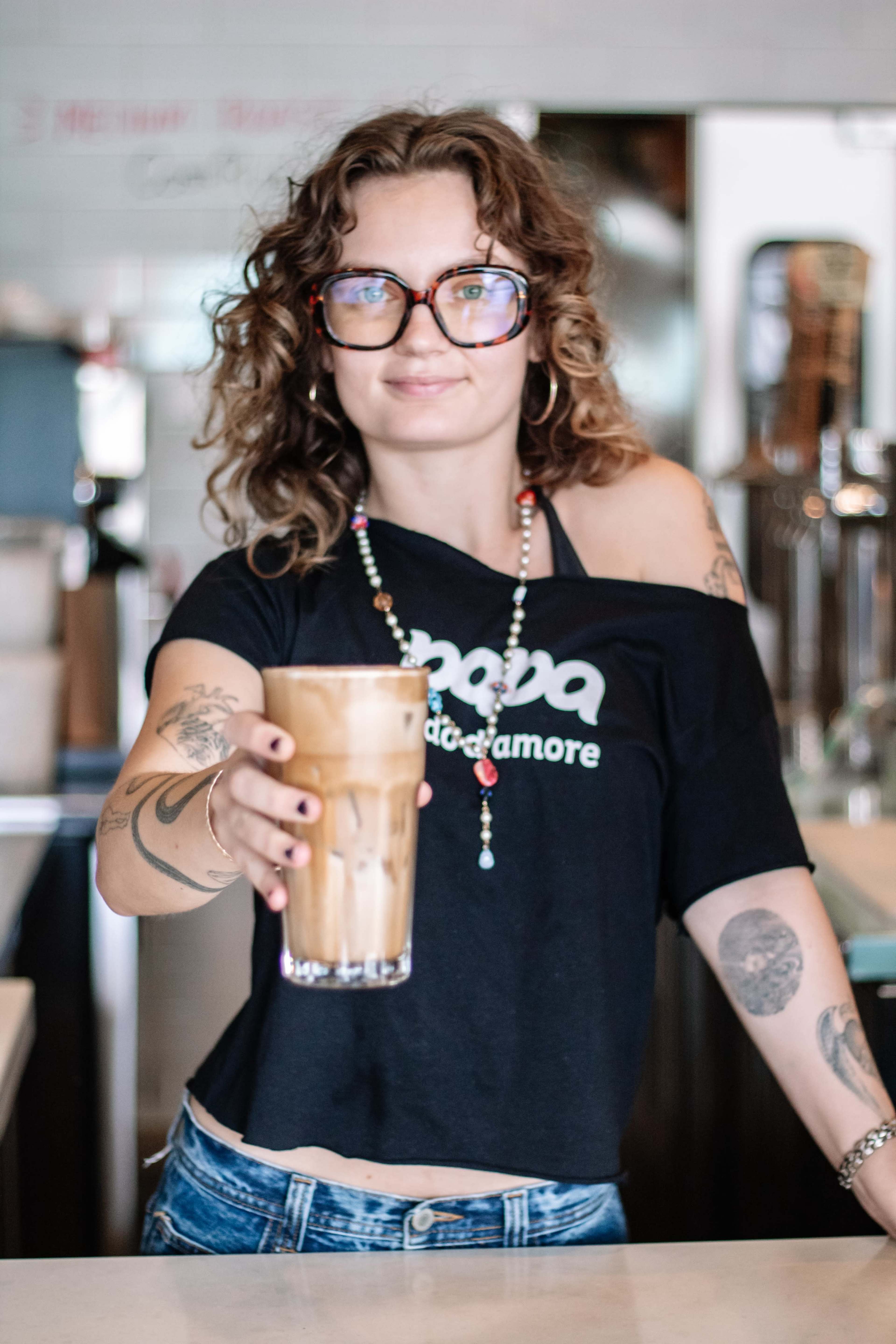 A barista with curly hair holds out a tall glass of iced coffee in a coffee shop setting.