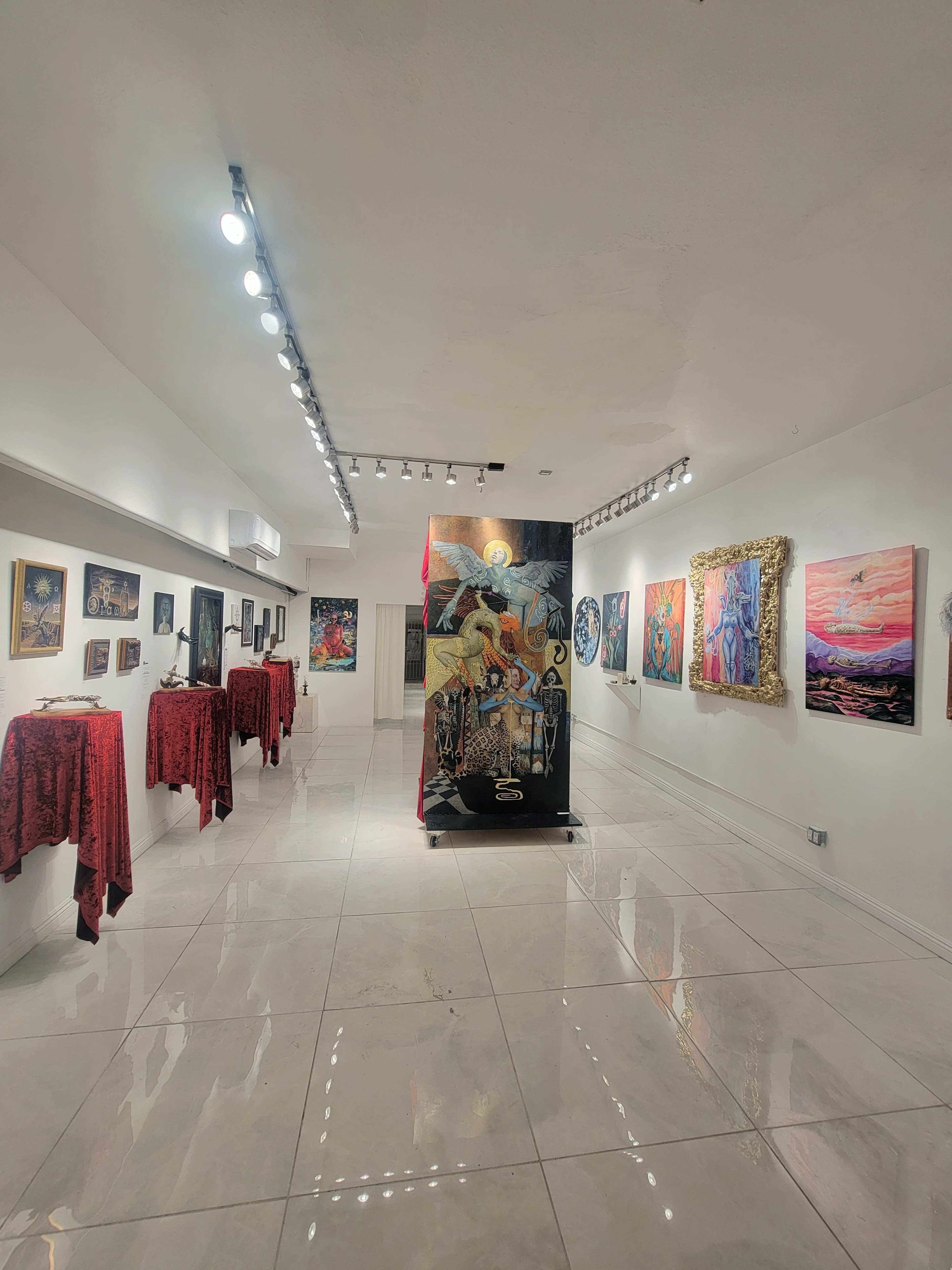 A gallery space with illuminated walls displaying various artworks, accompanied by tables covered in red cloths.
