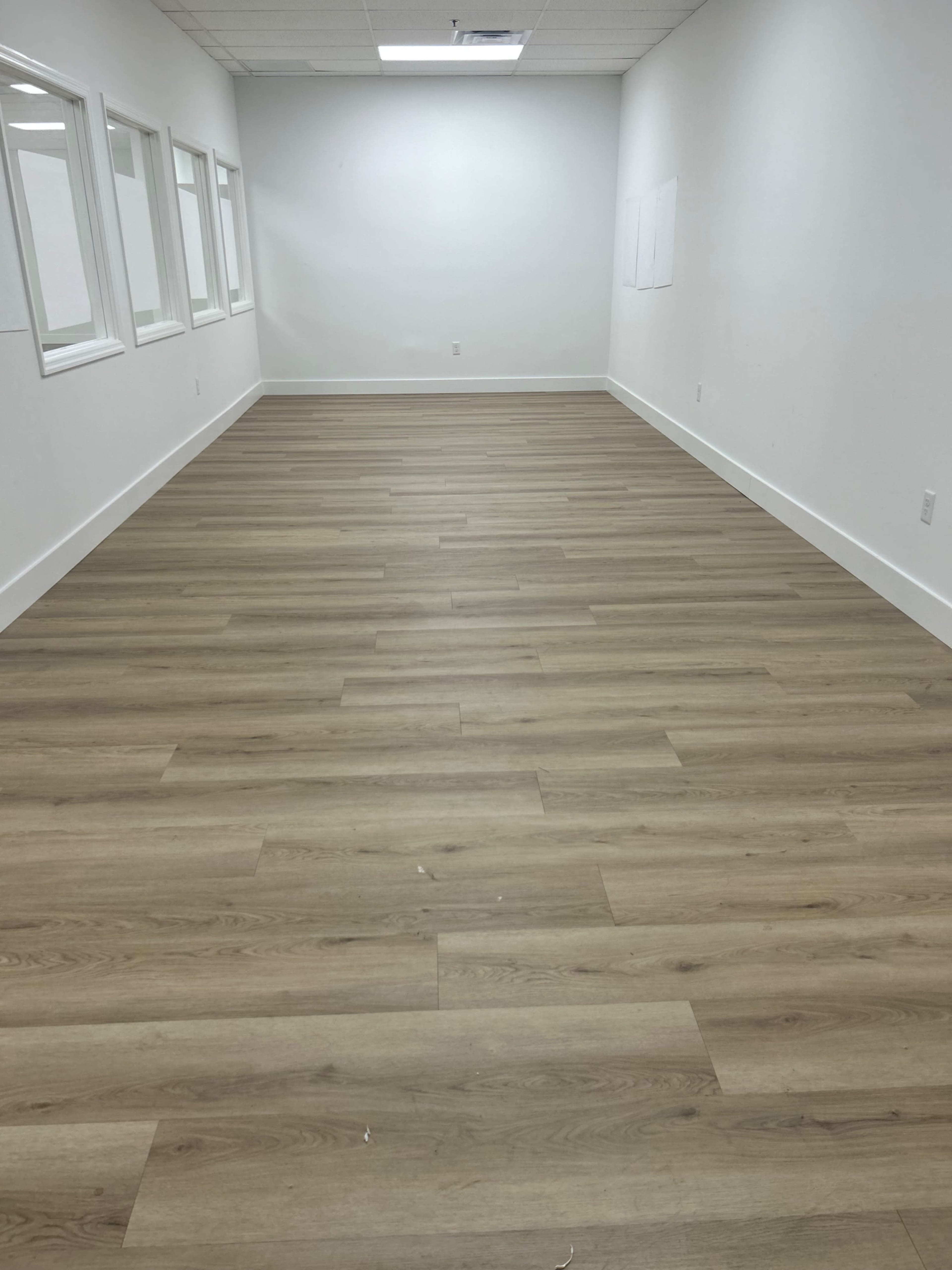 The image shows a vacant room with light wooden flooring and white walls, featuring several windows along one side.