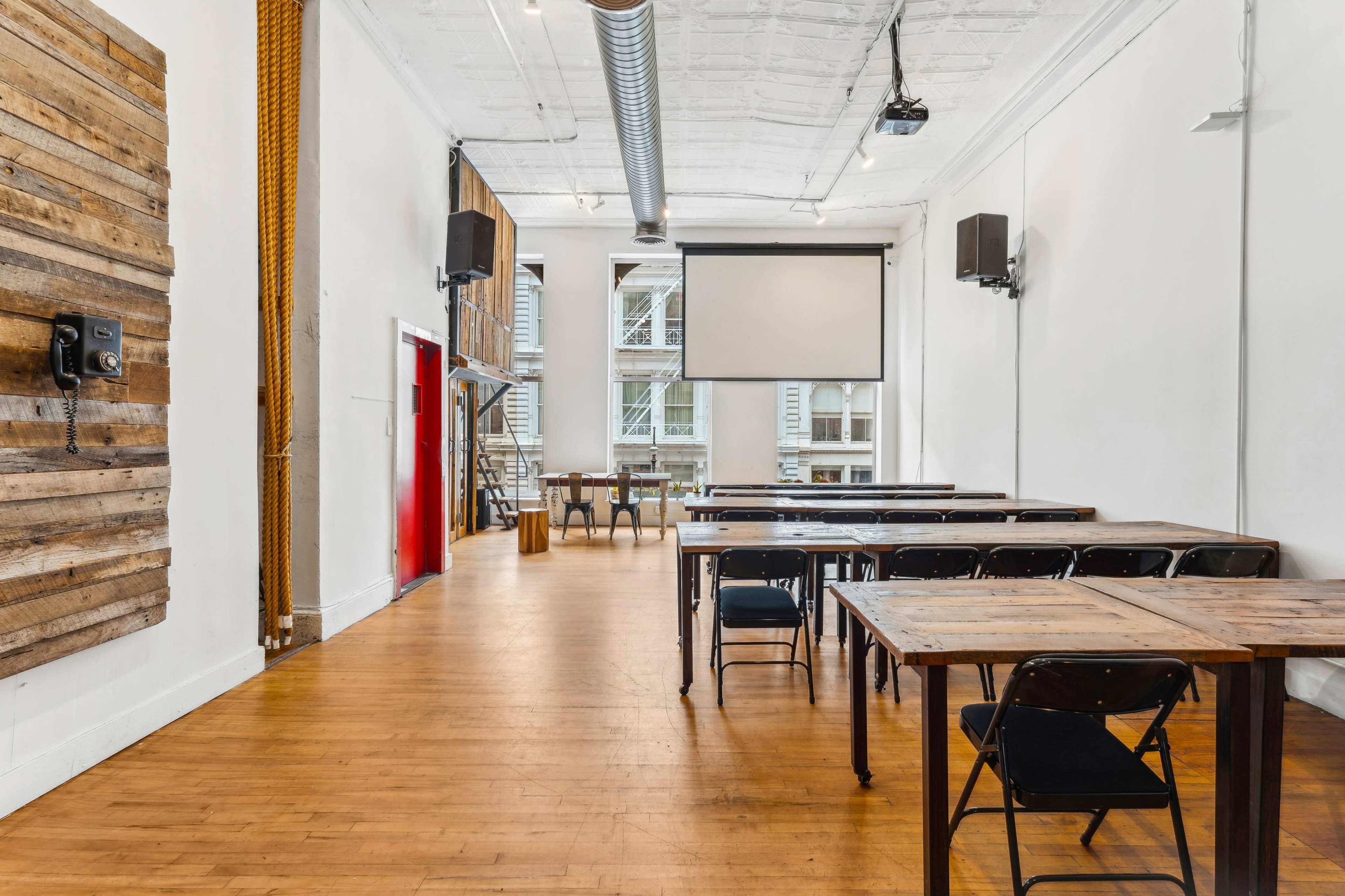 A spacious classroom with wooden tables and chairs arranged in a linear fashion, a projector screen mounted on the wall, and large windows allowing natural light to enter.