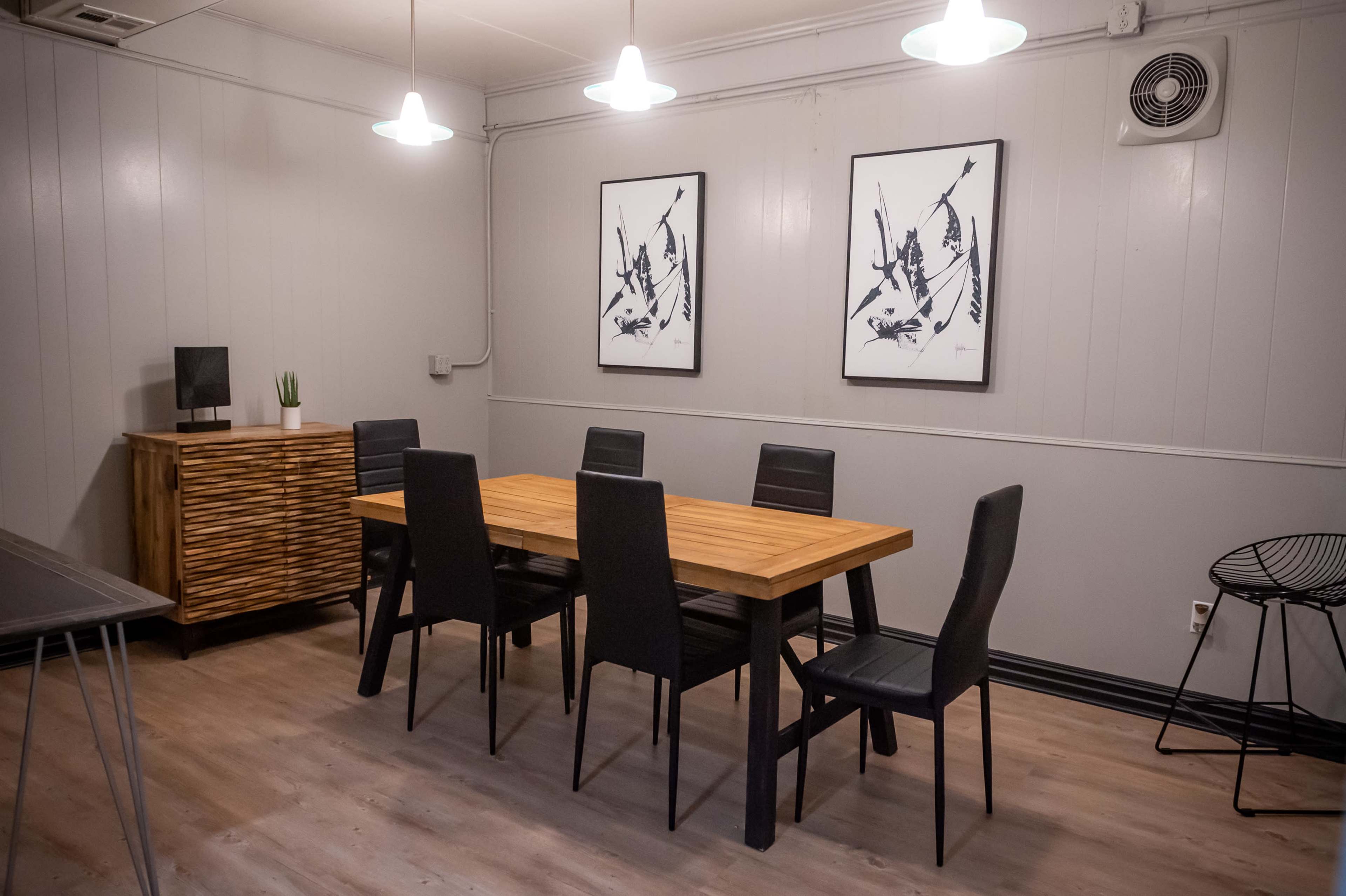 A dining area features a wooden table surrounded by black chairs, with two framed abstract artworks on the wall and a wooden cabinet nearby.