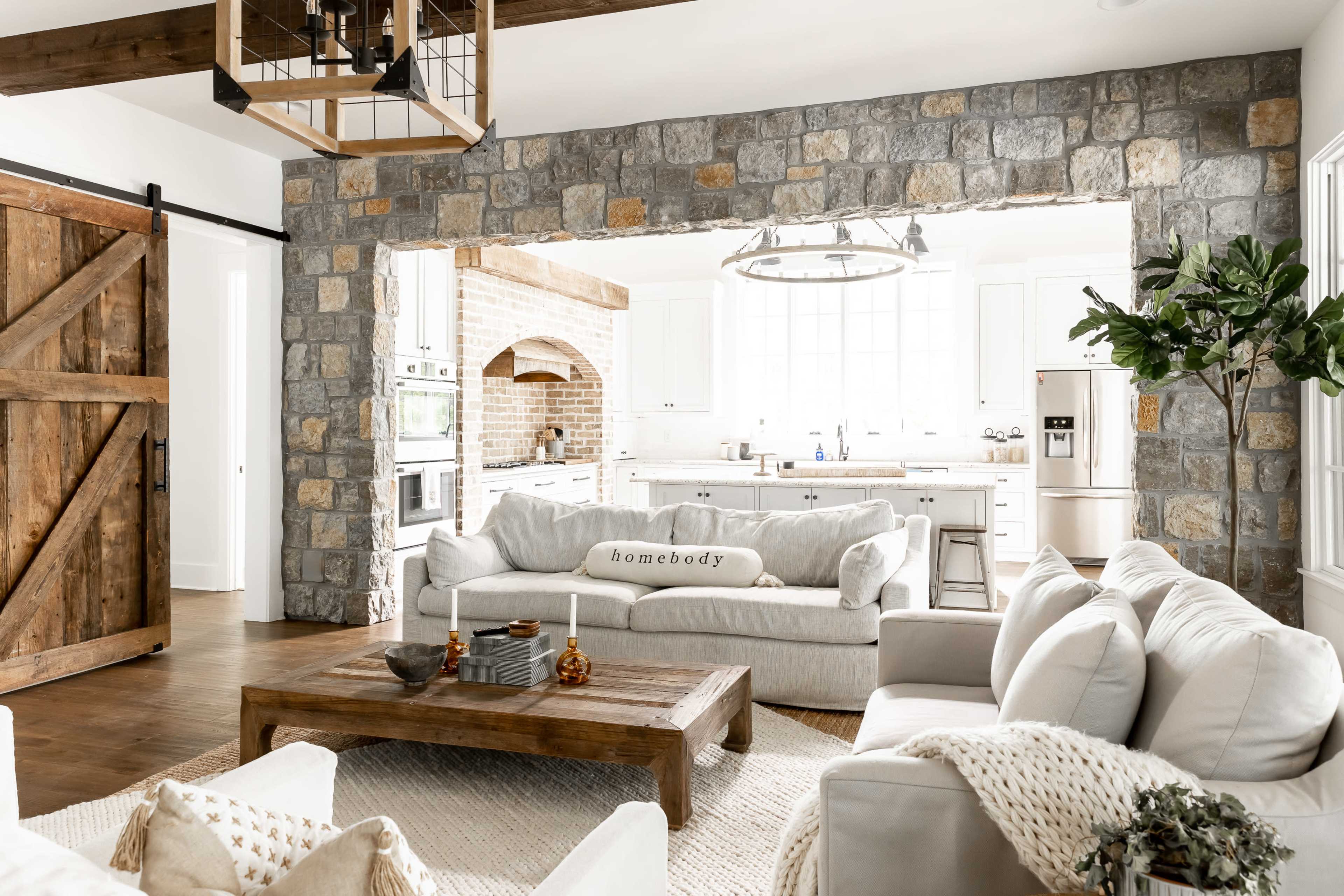 A spacious living room features a light gray sofa, a wooden coffee table, and a stone archway leading to a bright kitchen.