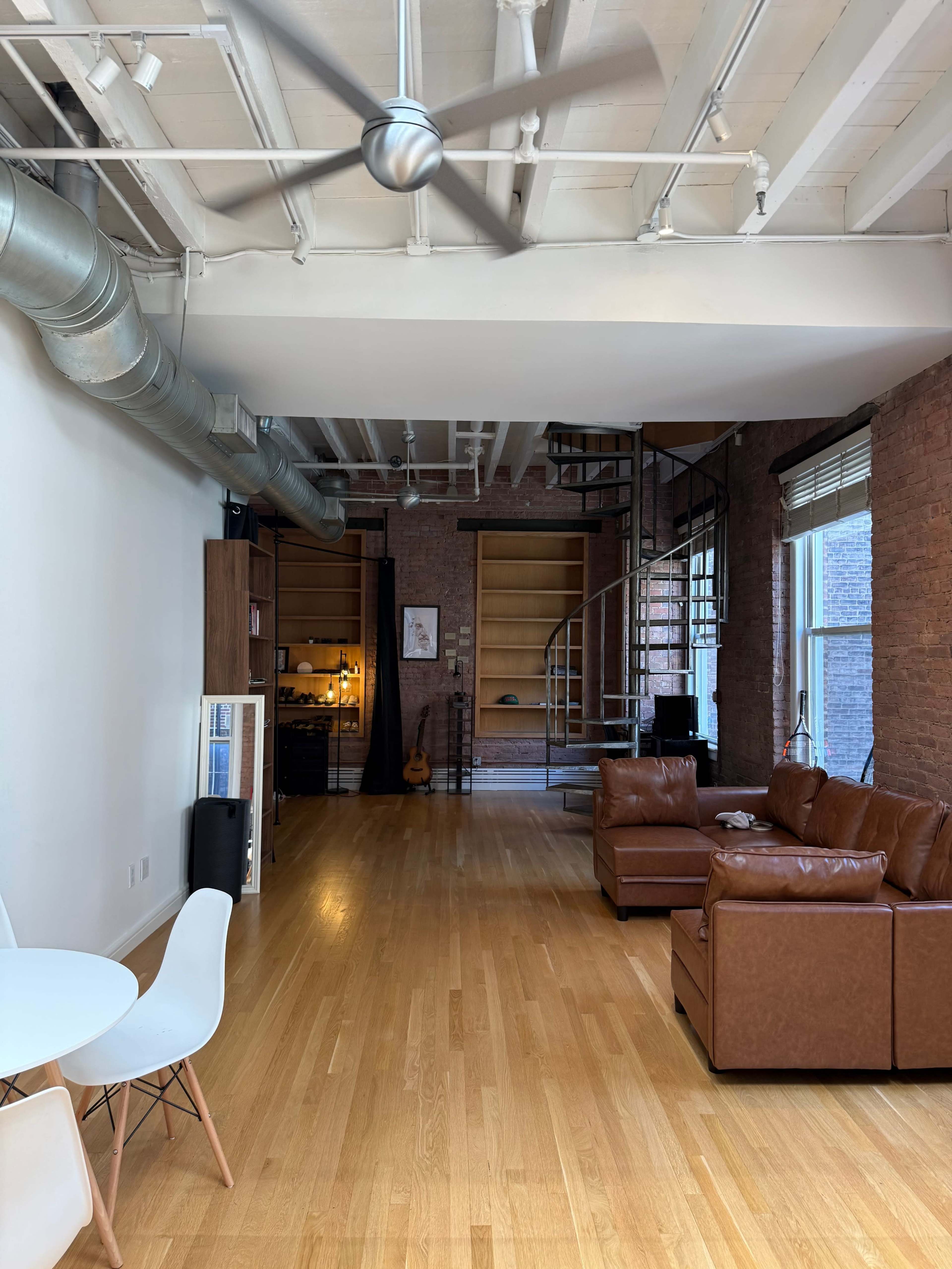 The image shows a spacious living area with a brown leather sectional couch, a wooden dining table with white chairs, and a spiral staircase leading to a lofted area.