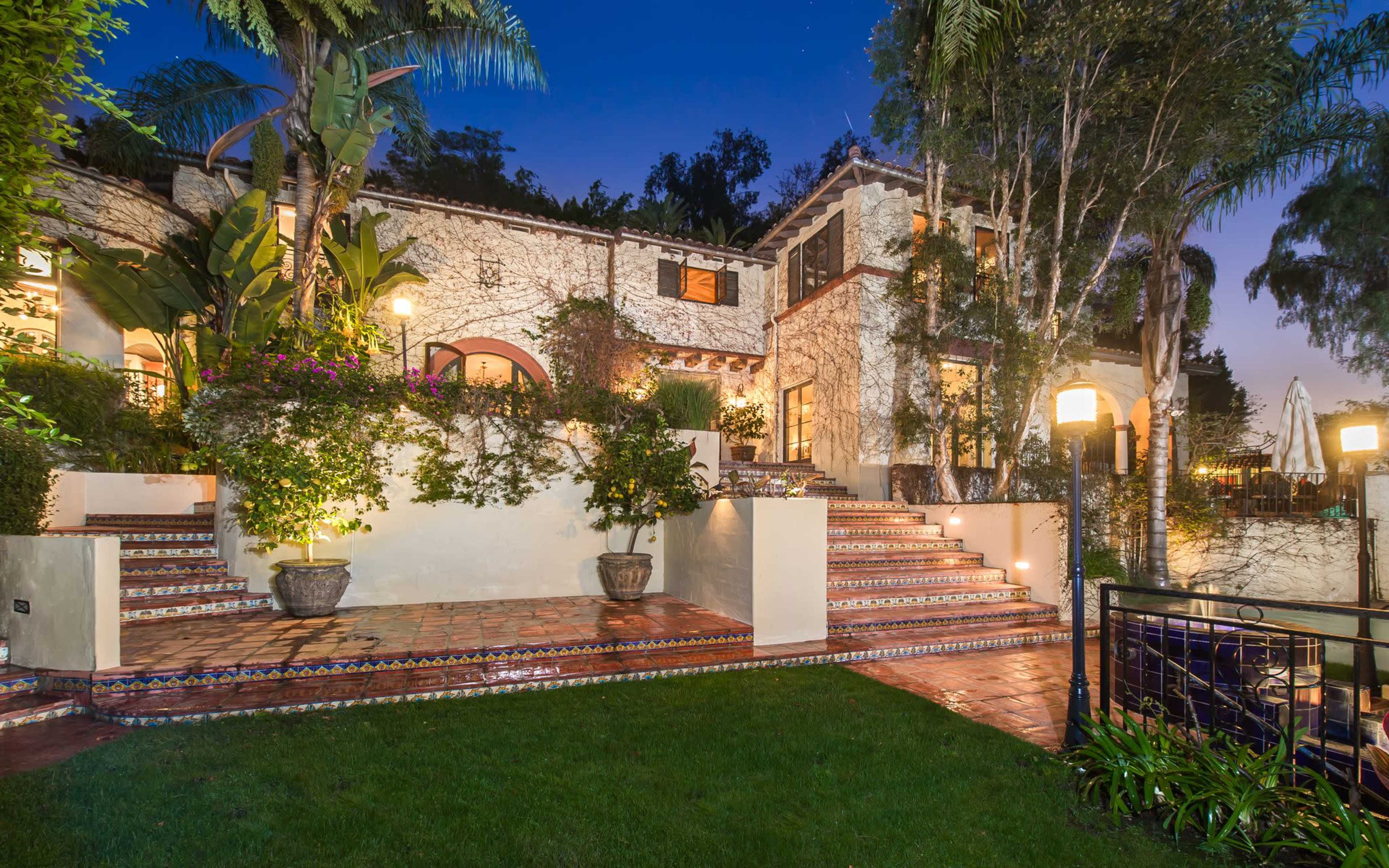 A two-story house with a terraced garden, surrounded by palm trees and illuminated by outdoor lighting.