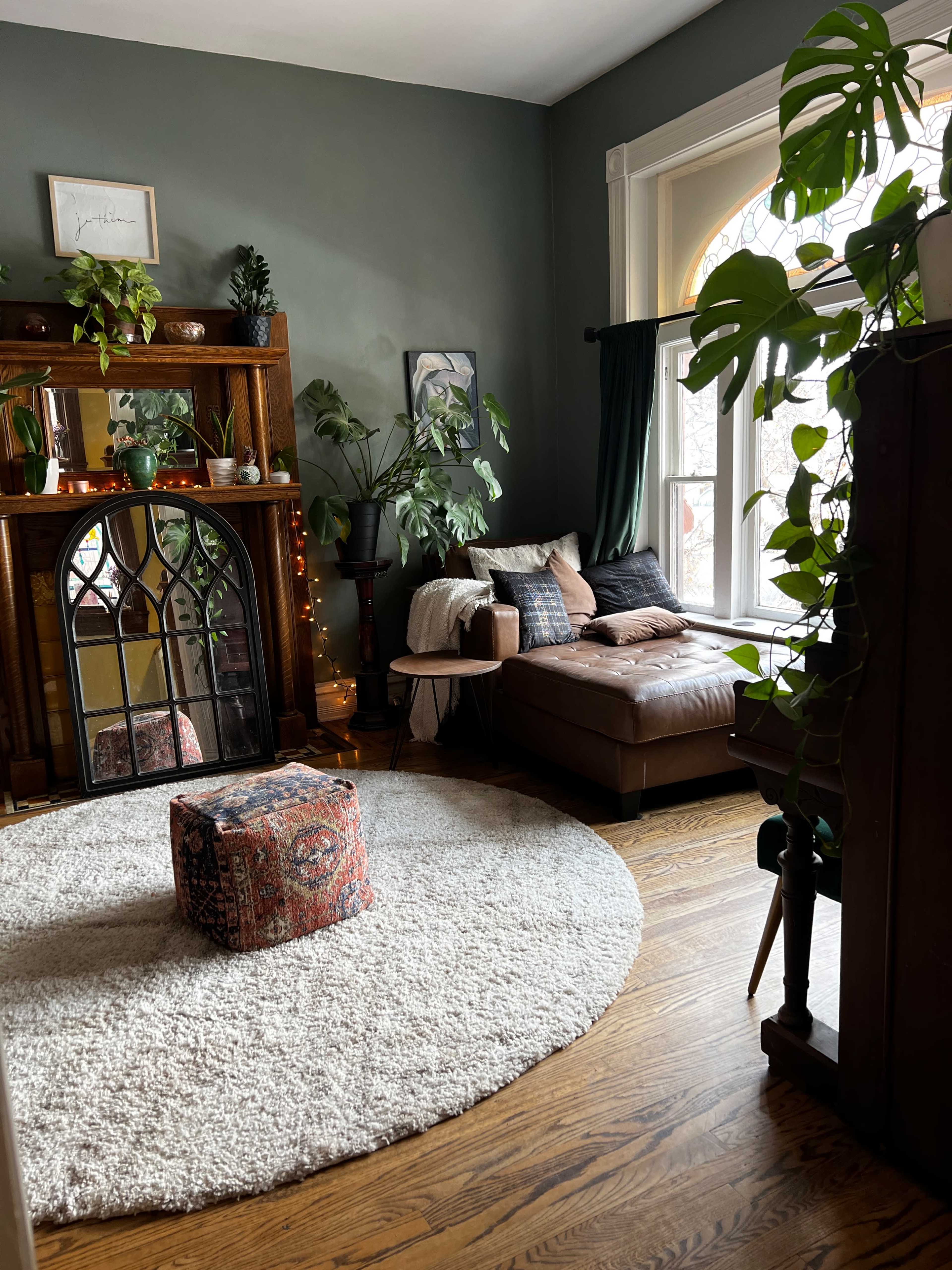 The image shows a cozy living room with a dark green wall, a large circular rug on the wooden floor, a mirrored fireplace, a brown sectional sofa, and several plants near a window.