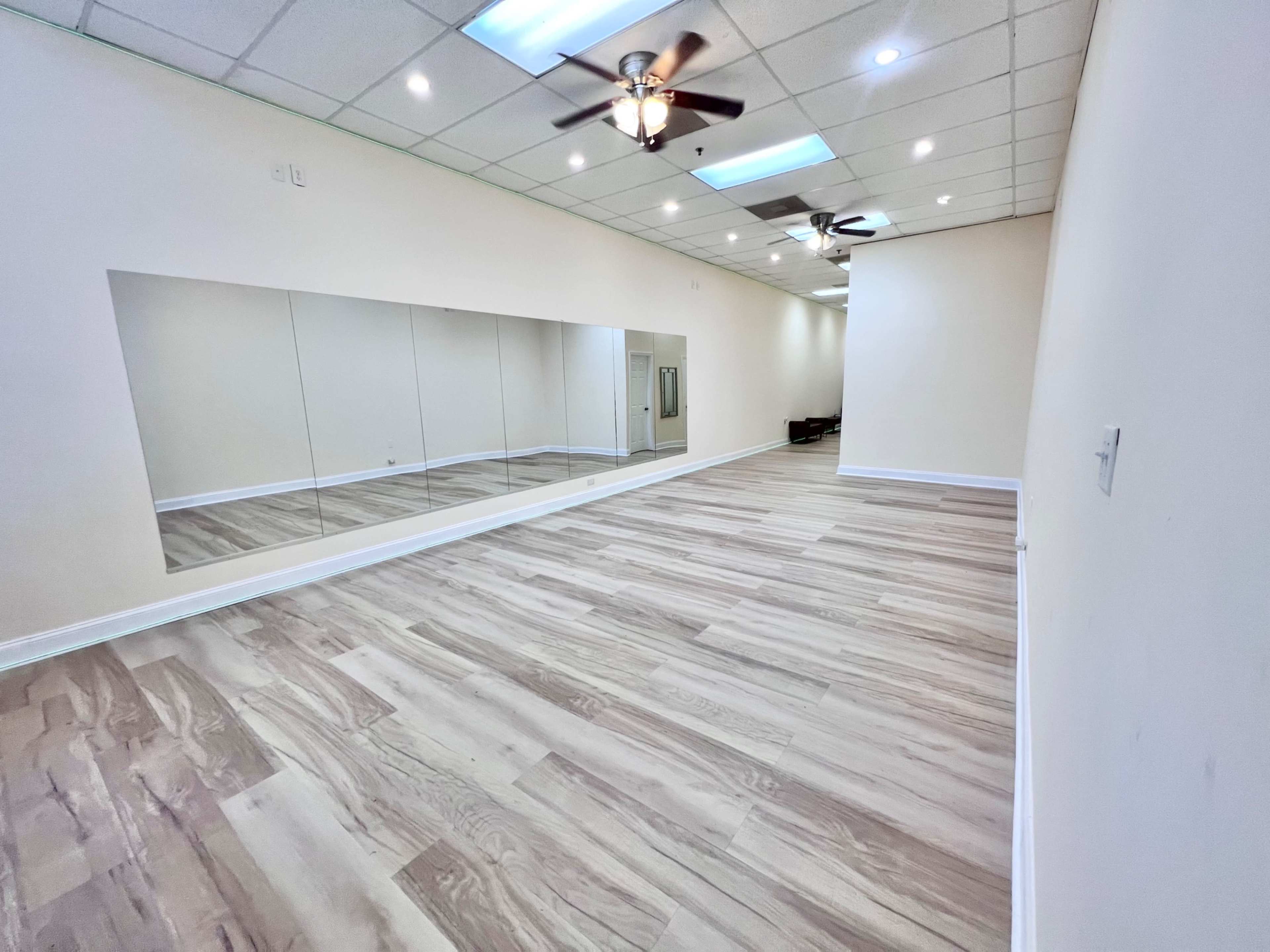 The image shows a spacious, well-lit room with a mirrored wall and wooden flooring.