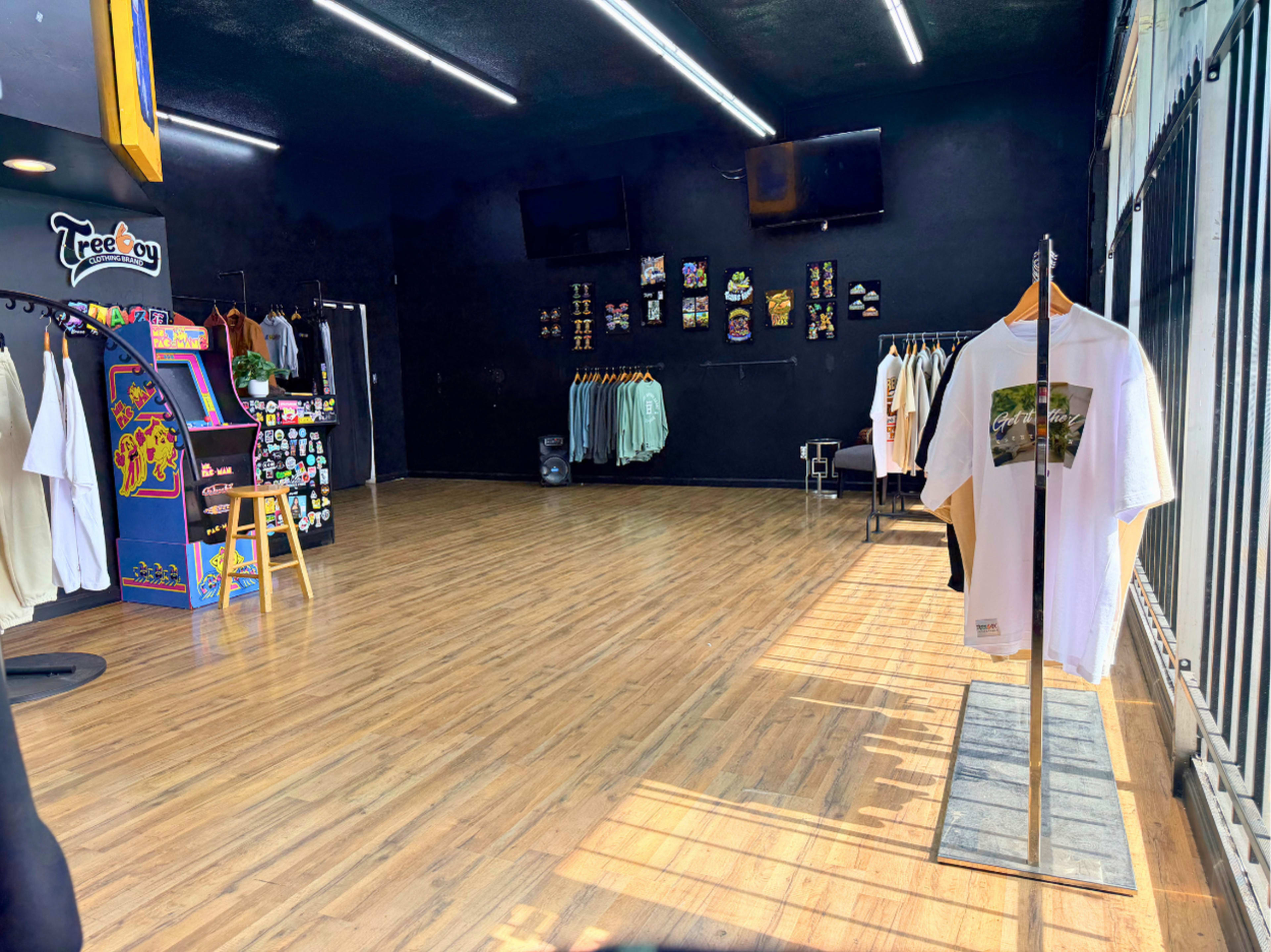 The image shows a retail clothing store with a wooden floor, featuring hanging garments on racks and an arcade game near the entrance.