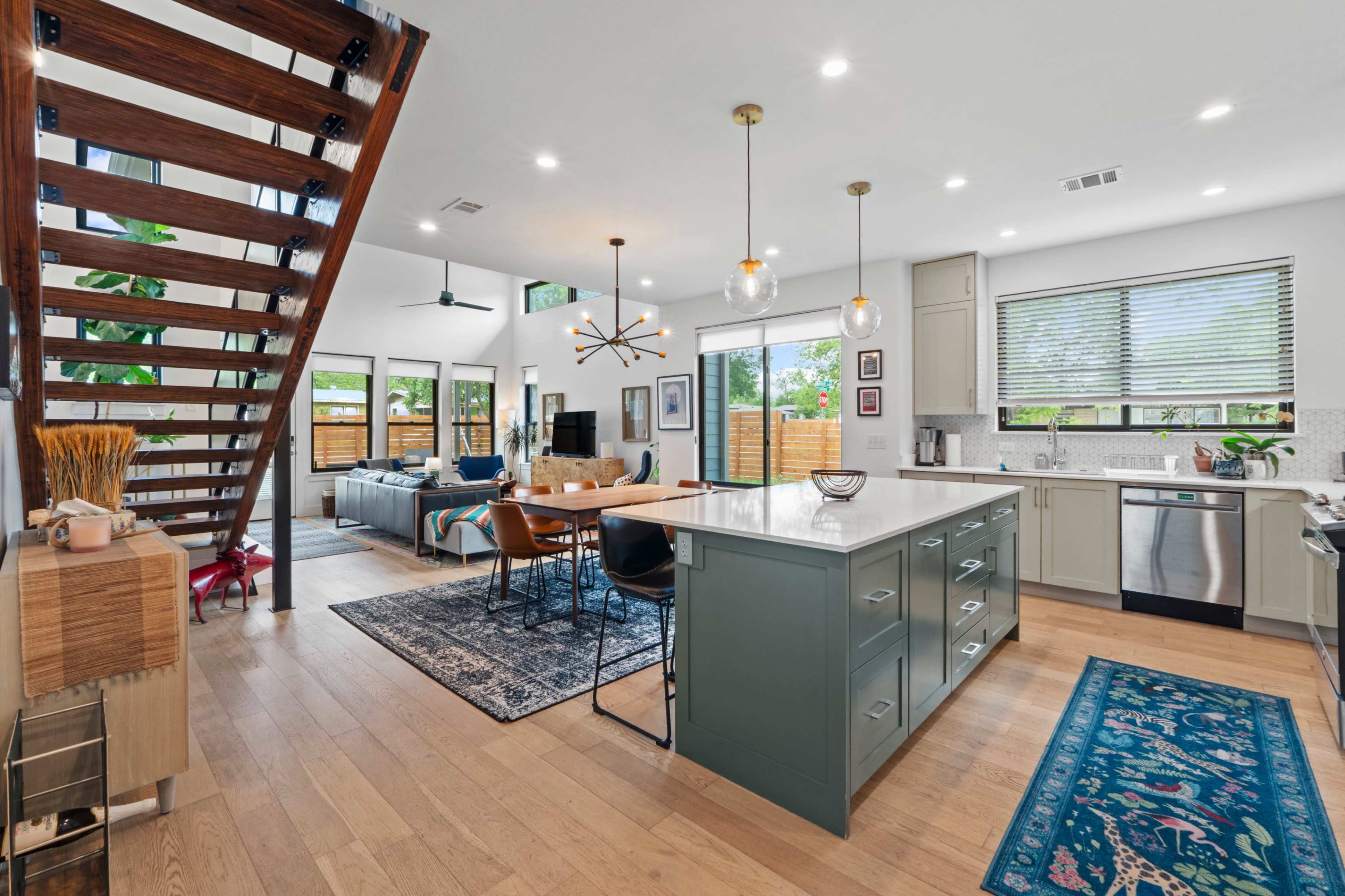 A modern kitchen and dining area feature an open layout with a central island, wooden staircase, and large windows that provide ample natural light.