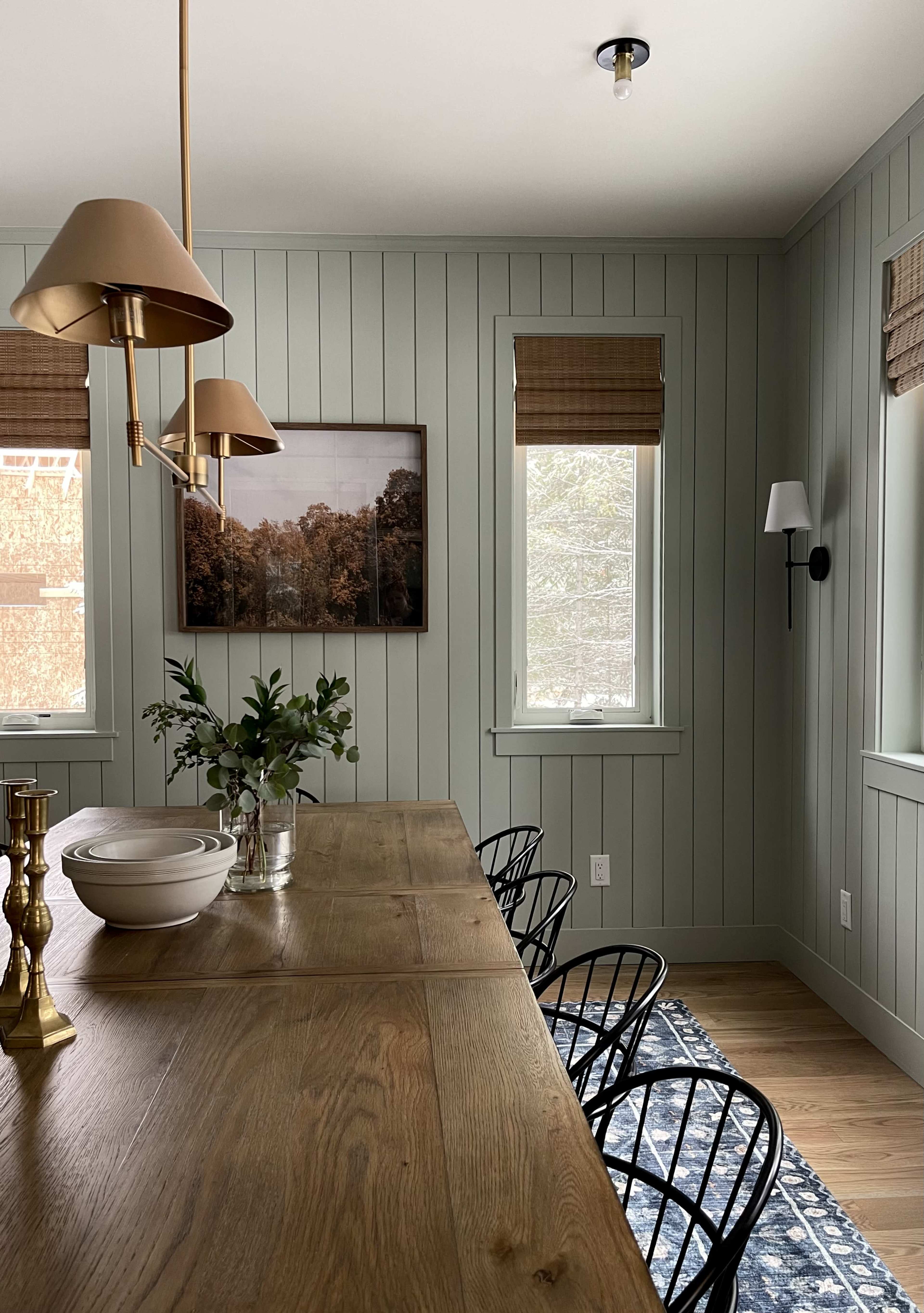 A dining area with a wooden table surrounded by black chairs, light fixtures overhead, and a large framed picture on the wall, complemented by natural light through two windows.